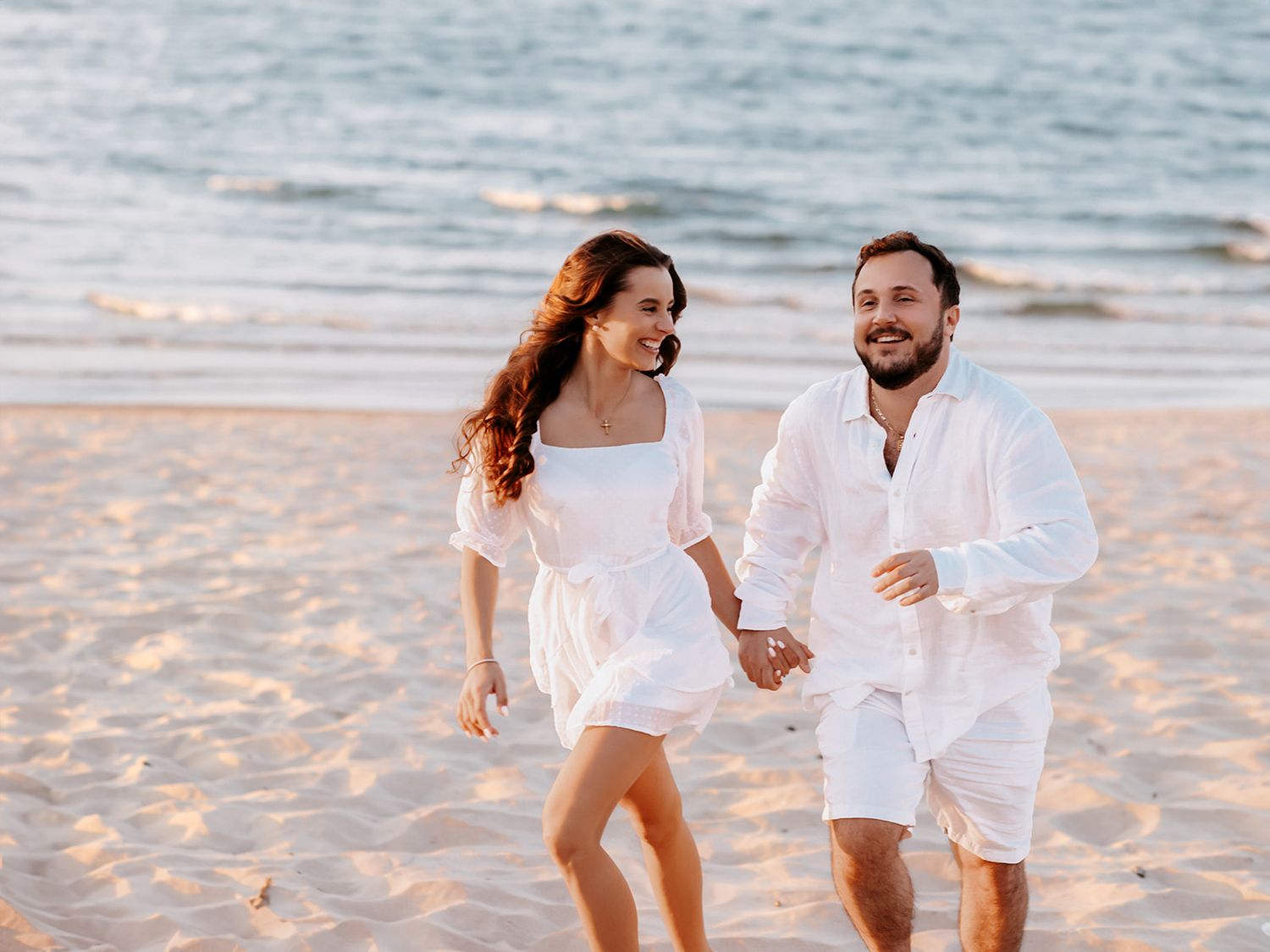 Joyful couple in white outfits running hand in hand along a sunny beach by the ocean.