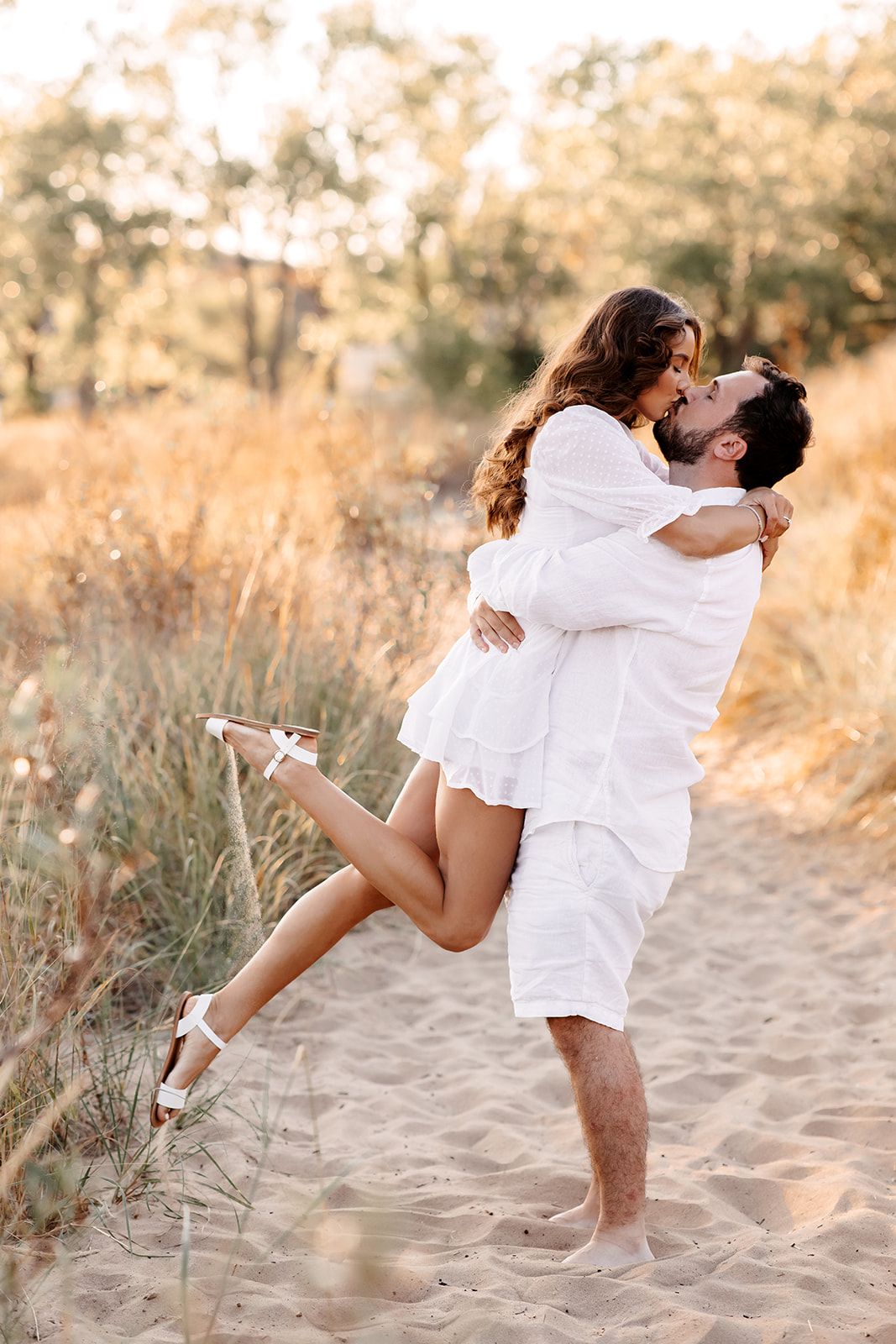 A couple in white outfits share a romantic kiss on a sandy path, the man lifting the woman.