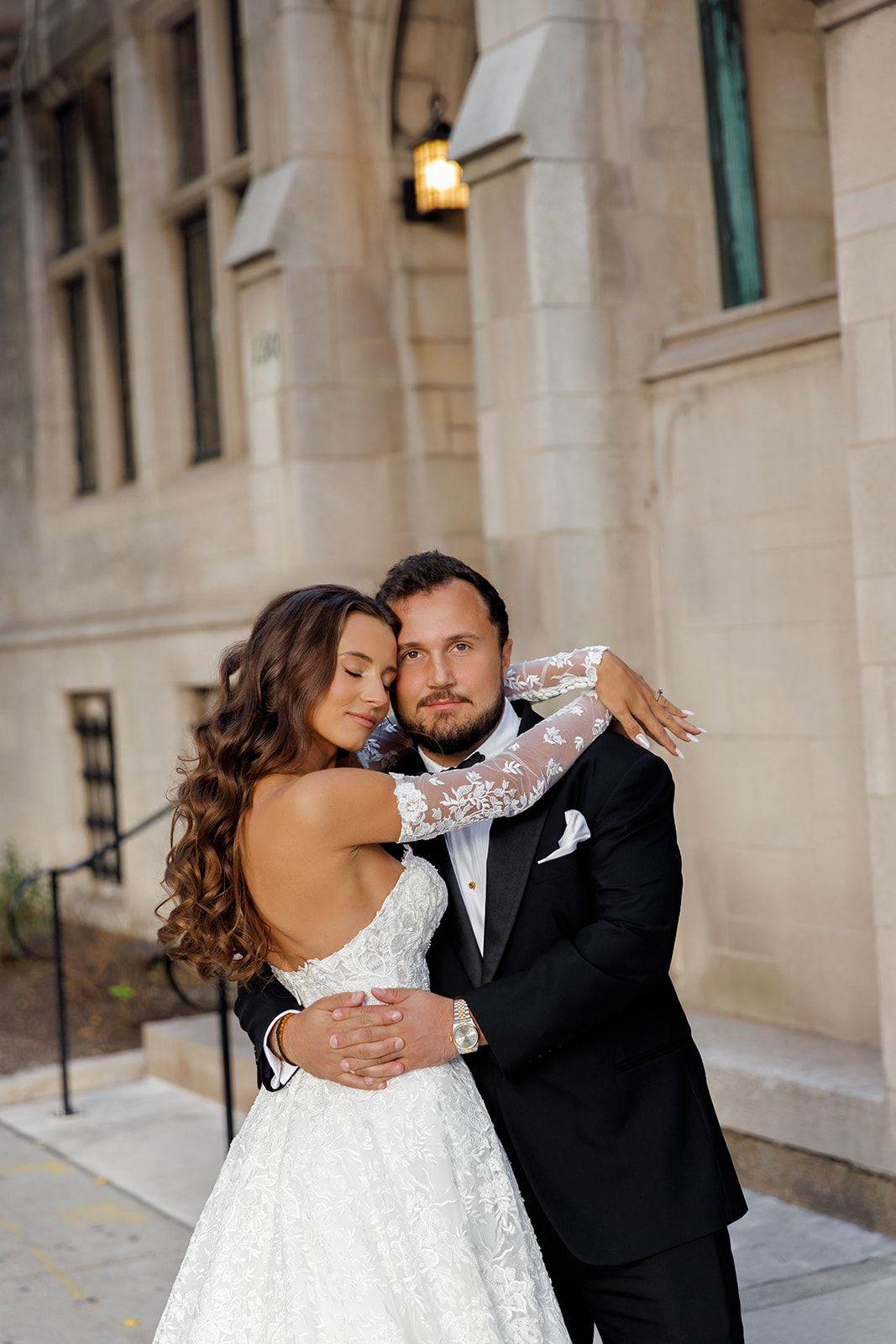 A bride in a backless lace gown embraces her groom in a black tuxedo outside a stone building.