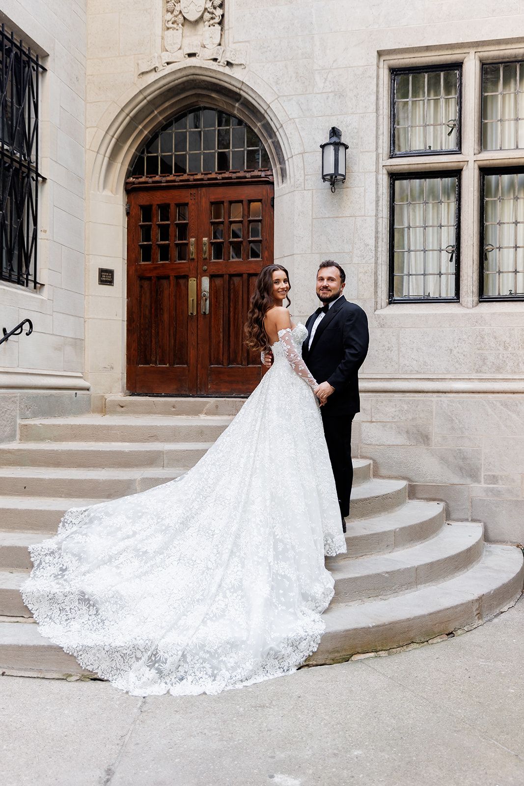 Bride in lace ball gown with long train and groom in tuxedo posing on stone steps before ornate wooden doors.