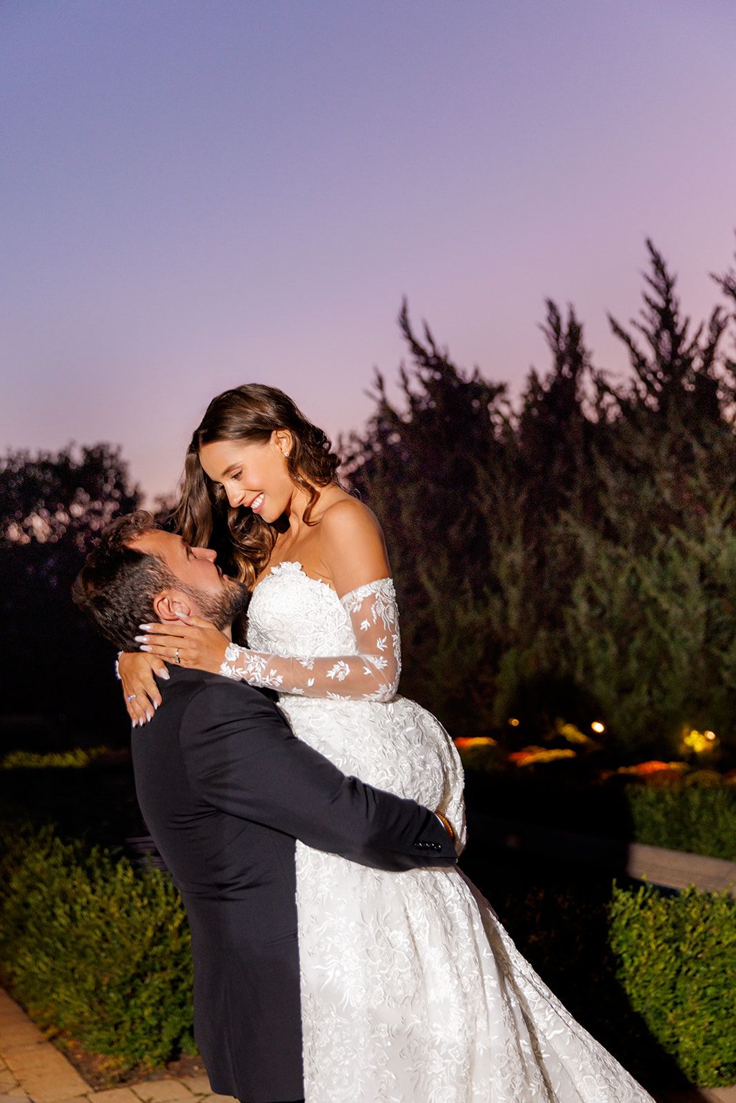 Groom lifts smiling bride in lace off-shoulder wedding gown at twilight against a purple sky backdrop.