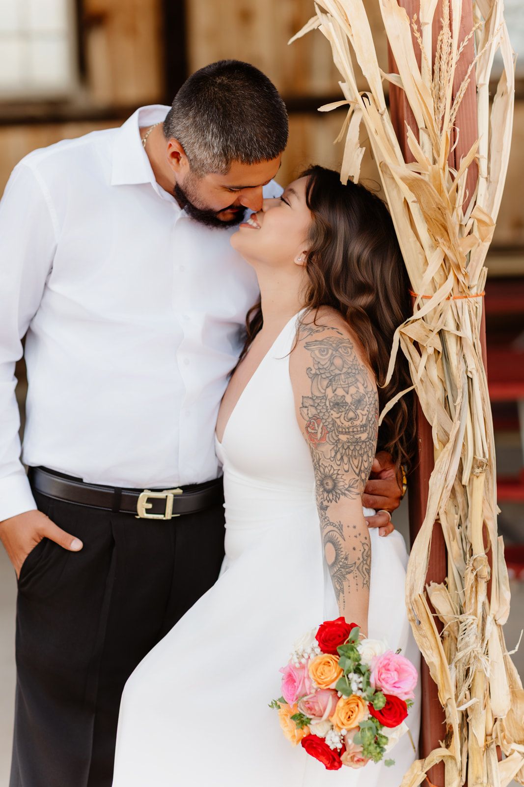 Bride and groom kissing near dried corn stalks, bride holding colorful bouquet.