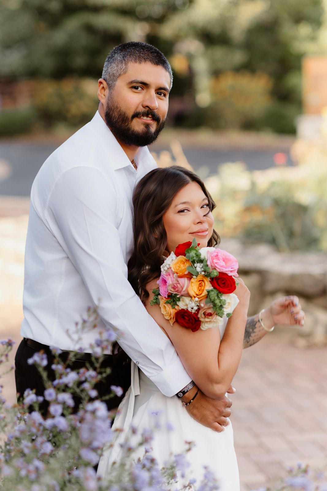A bride holding a colorful bouquet embraces with her bearded groom in white shirt outdoors at golden hour.