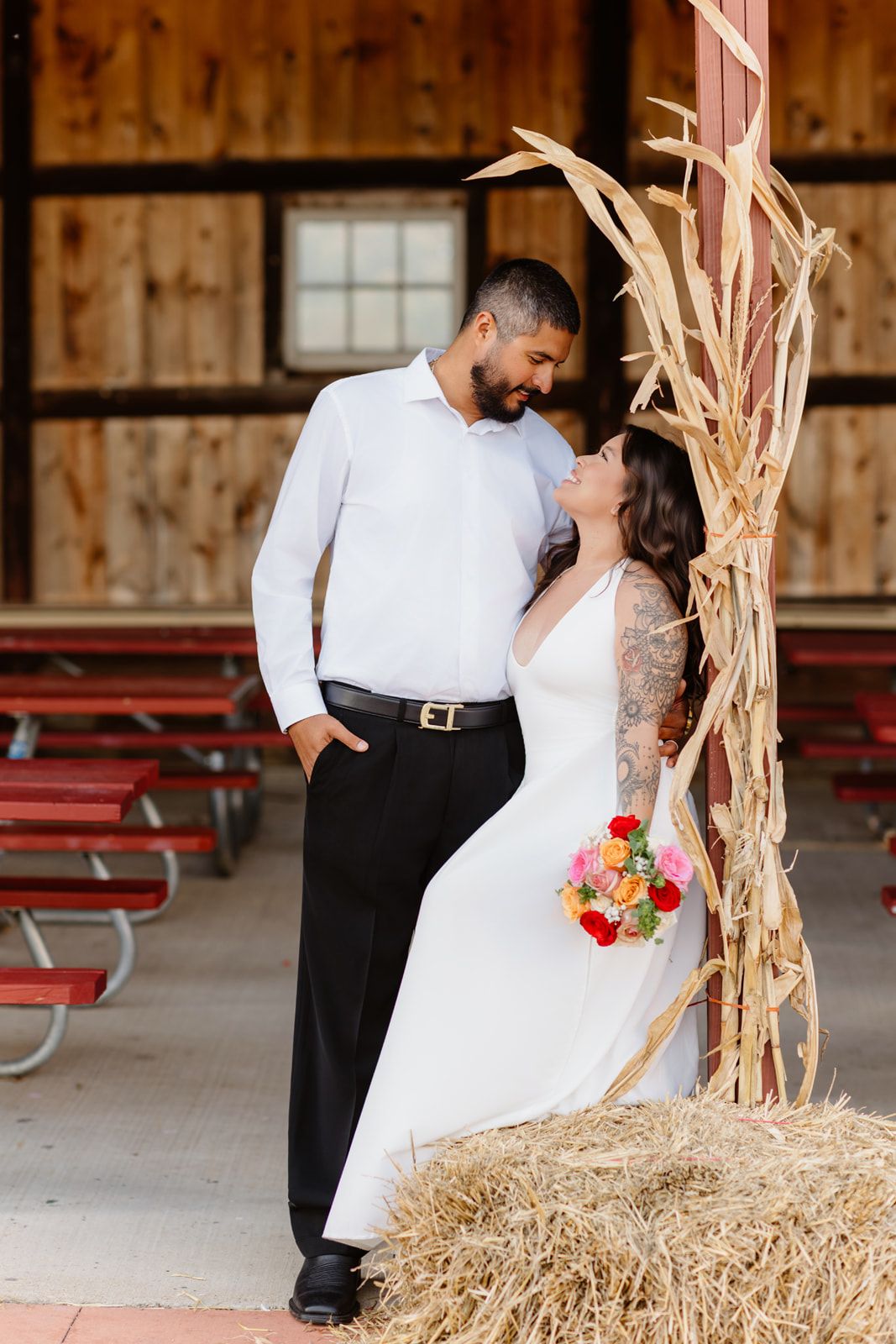 Bride and groom share a romantic moment beside a hay bale and dried corn stalks in a rustic barn wedding venue.