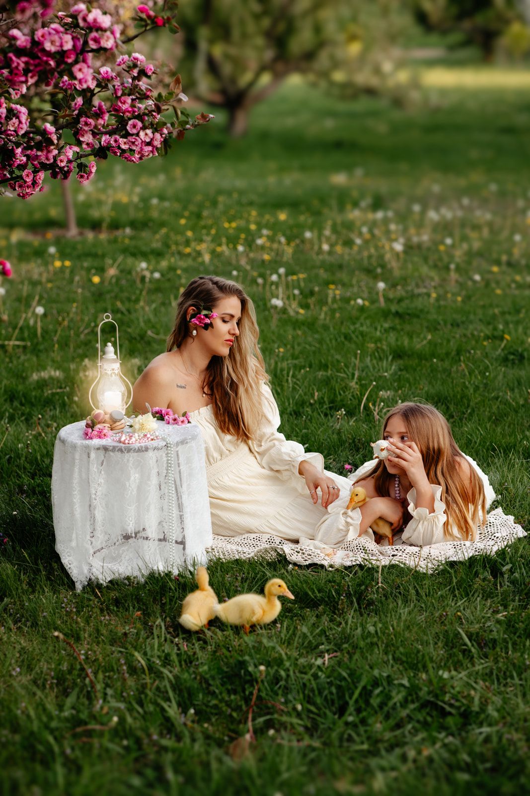 Mother and daughter in white dresses enjoy a spring picnic with ducklings under blooming pink trees on green grass.