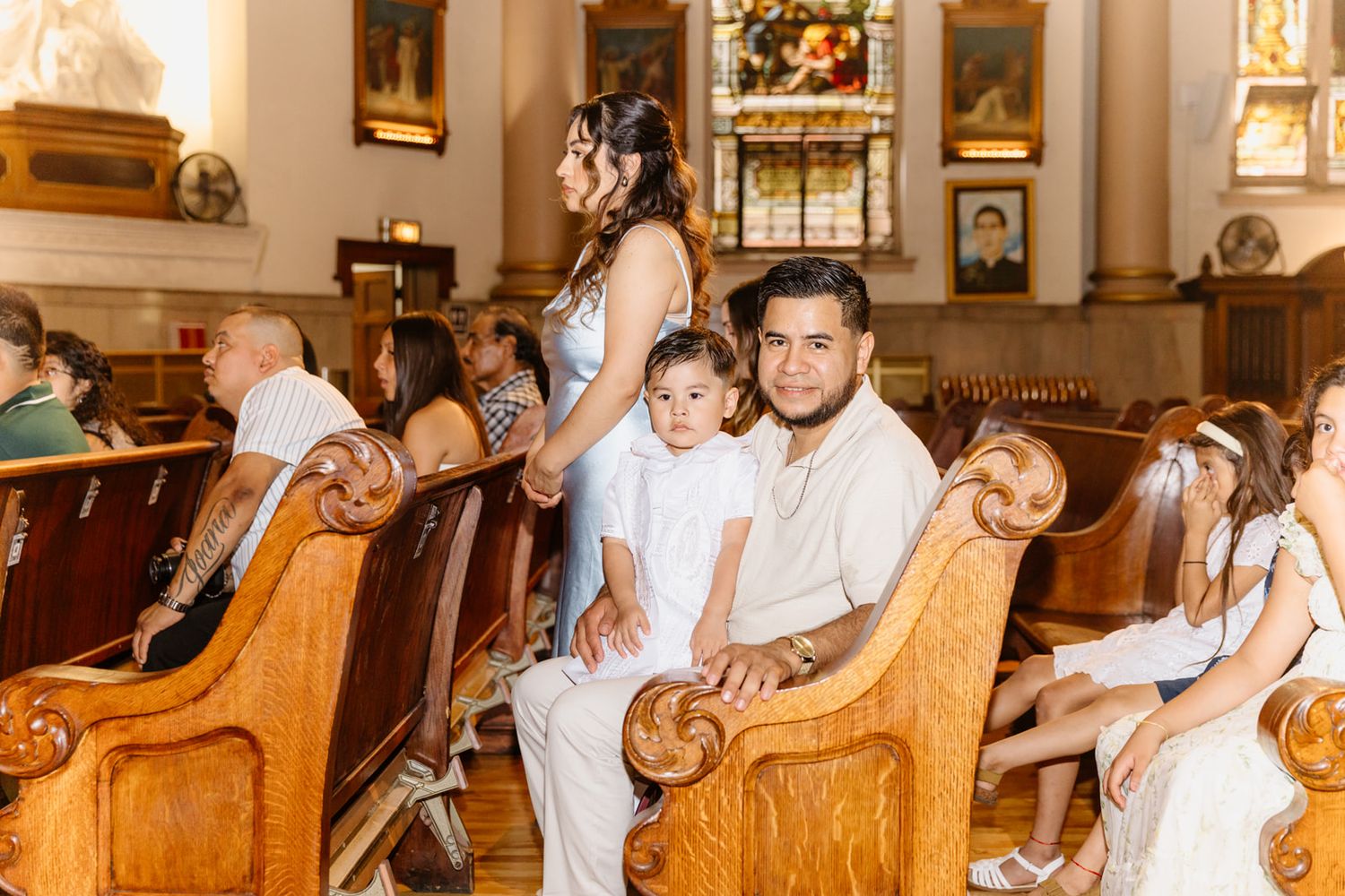 Family dressed in white sitting in wooden church pews during a religious ceremony with stained glass windows in background.