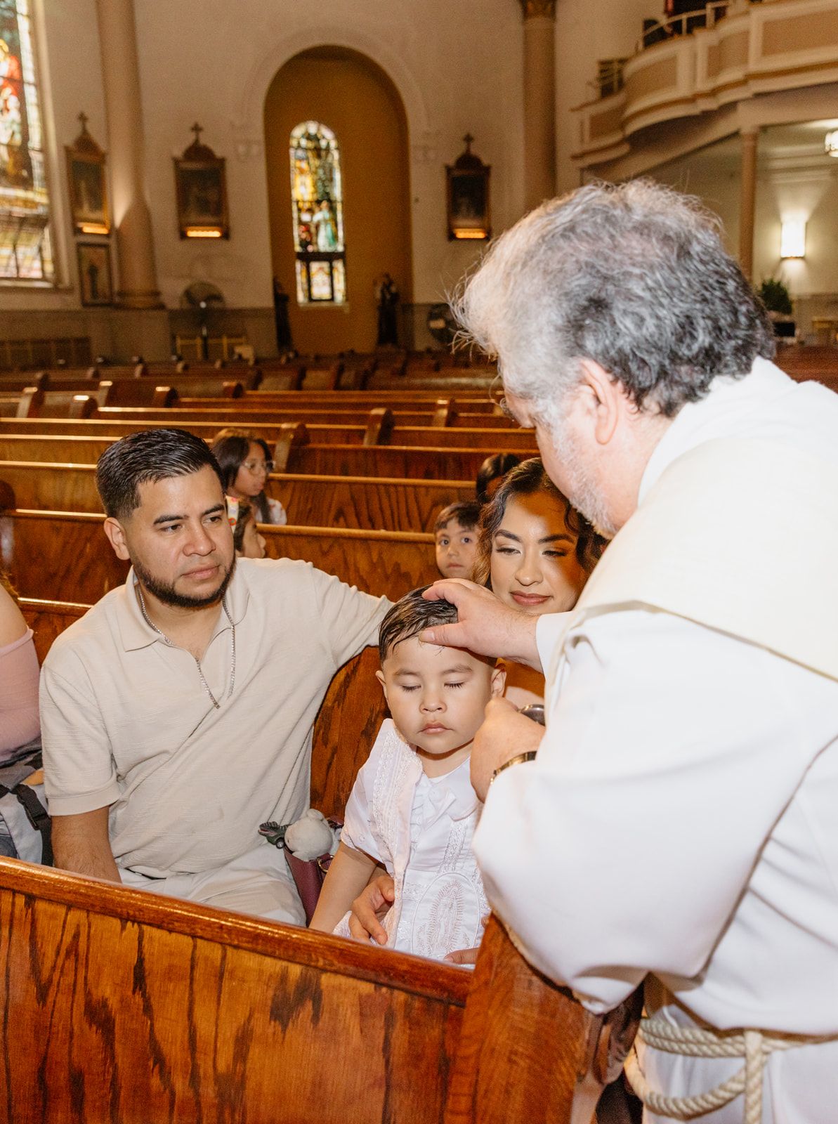 Catholic priest blesses young child during baptism ceremony with parents seated in church pew.