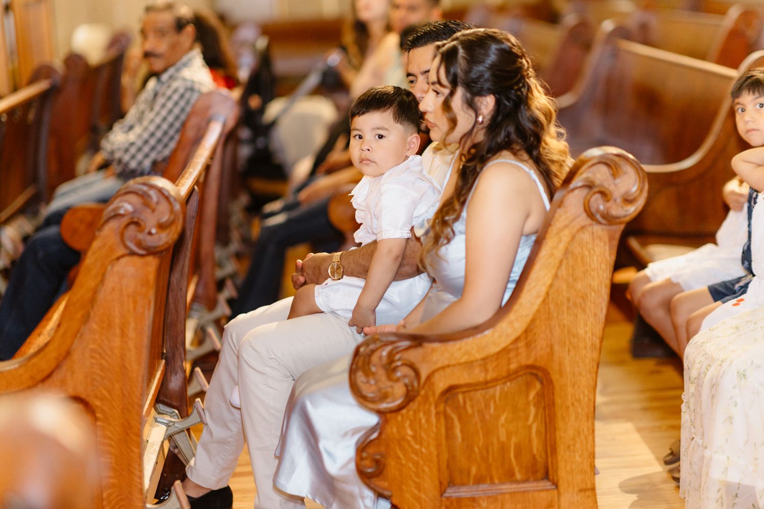 Young mother holds her baby dressed in white while seated in wooden church pews during a baptism.