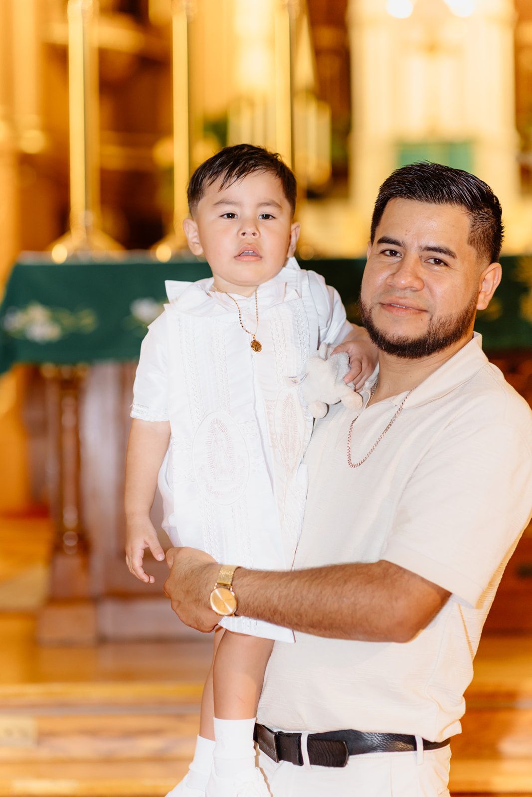 Father holding young child in white outfit inside ornate church during baptism ceremony.