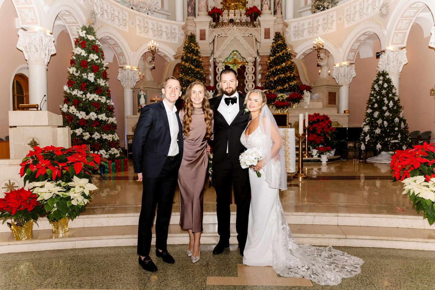 Wedding couple and two guests smiling in Christmas-decorated church with poinsettias and holiday trees.