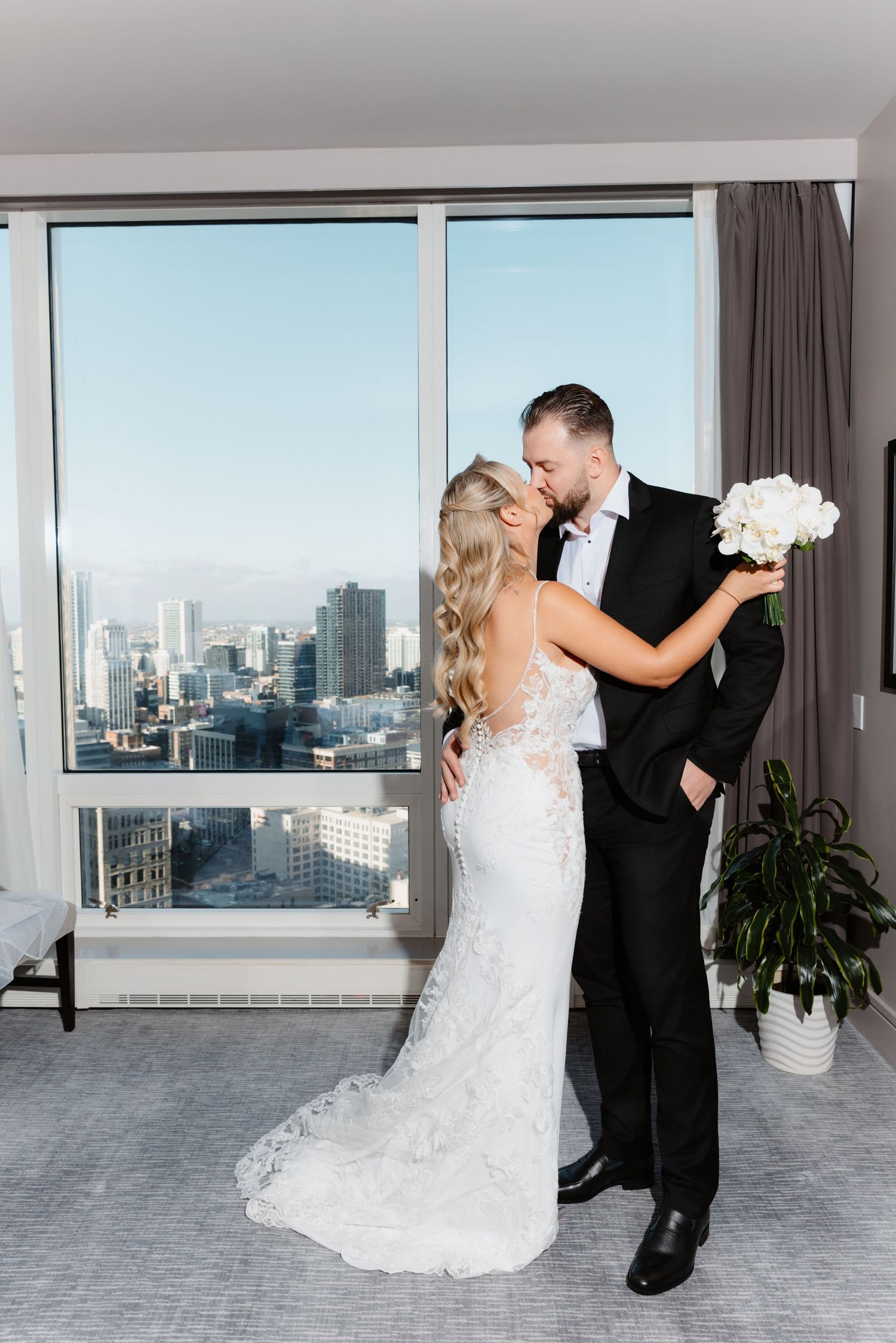 Bride and groom share a romantic kiss holding white bouquet with urban skyline in background.