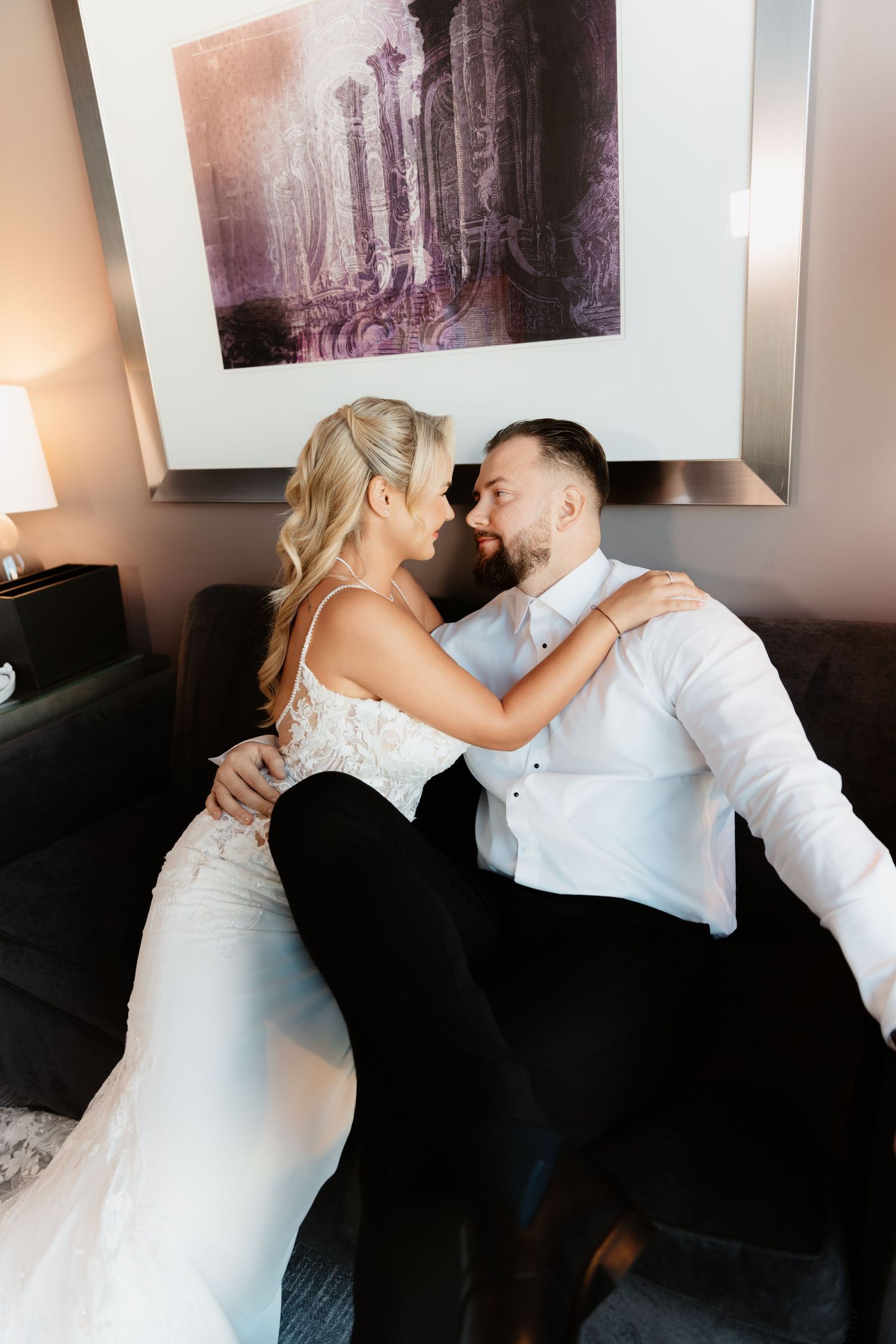 Bride in lace wedding gown and groom share intimate moment on sofa in hotel suite.