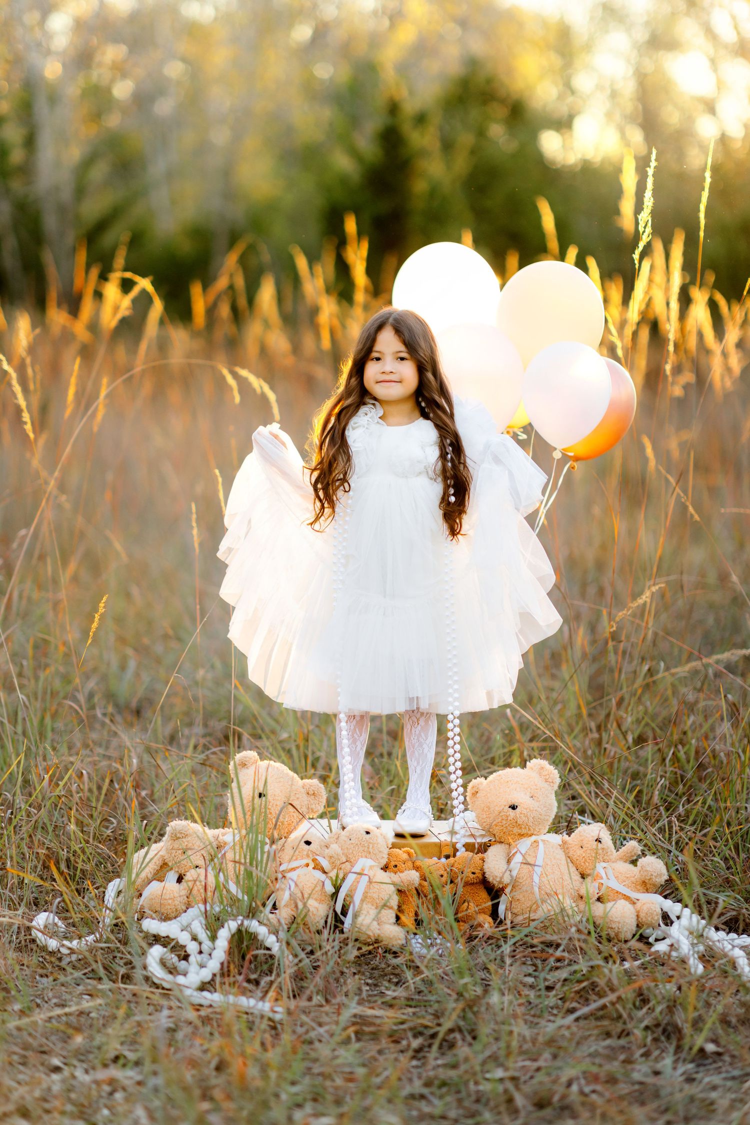 Young girl in white dress with angel wings holds balloons standing on suitcase with teddy bears in golden field.