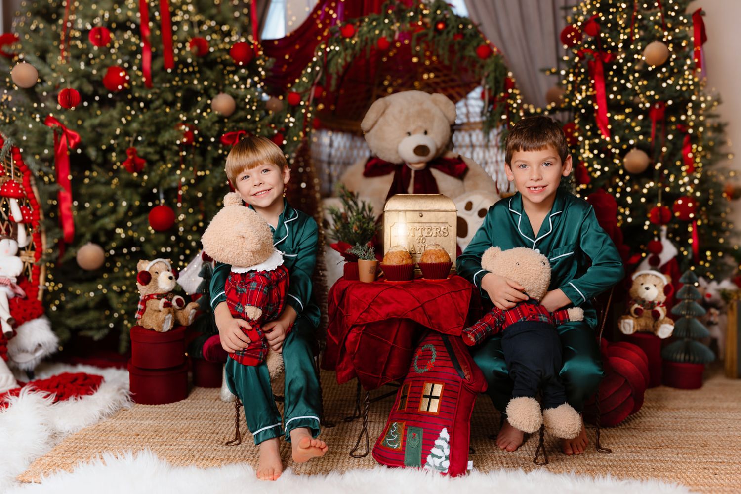 Two children in green pajamas sit among Christmas trees, teddy bears, and festive decorations in a cozy holiday photo.