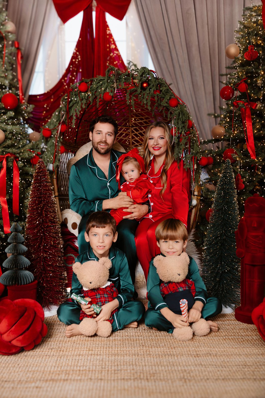 Family of five in red and green pajamas posing with teddy bears beside decorated Christmas trees and festive holiday decor.