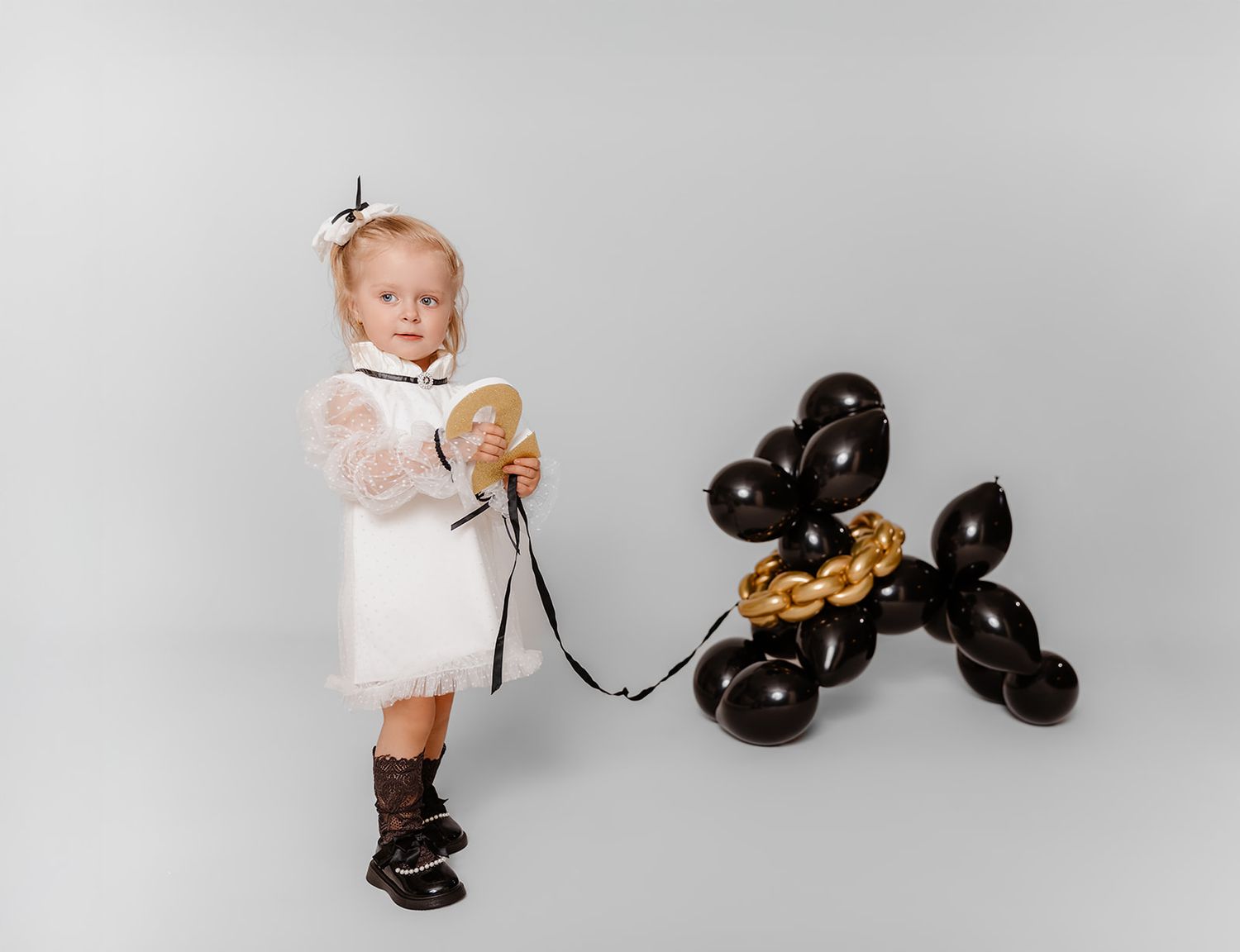 A toddler girl in a white dress holds a leash attached to a black balloon dog sculpture on a gray background.