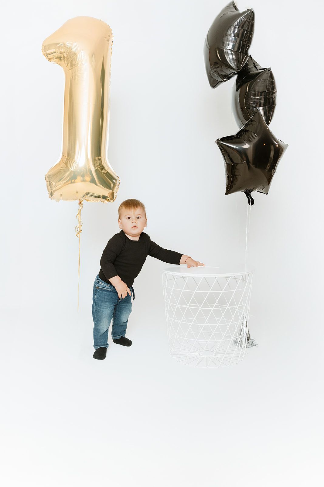 Toddler in black top and jeans stands by white basket with gold number one and dark star balloons.