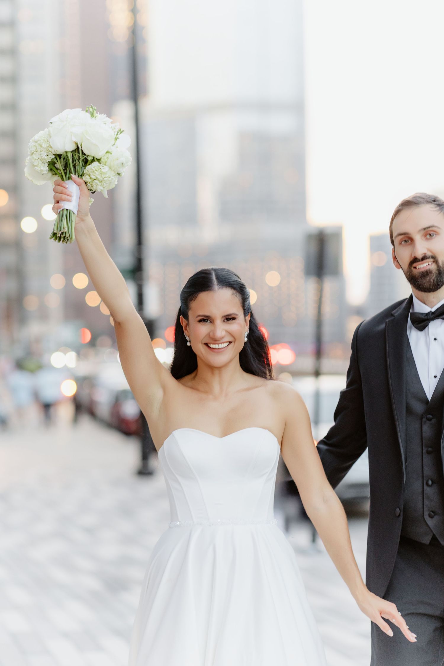 Joyful bride raises white bouquet in celebration while walking with groom in black tuxedo on a city street.