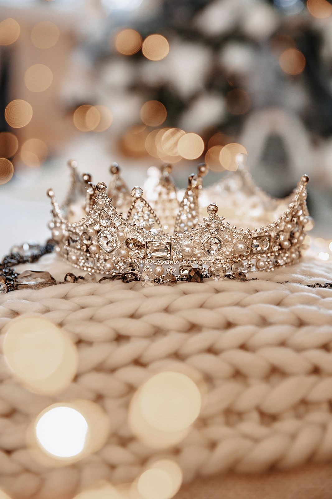 Ornate silver crown adorned with crystals and pearls resting on a white knit surface with bokeh lights in the background.