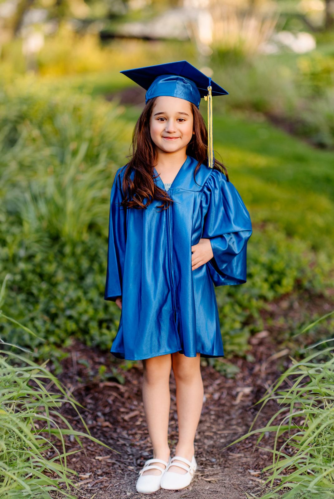 Young girl in shiny blue graduation cap and gown posing on garden path surrounded by greenery.