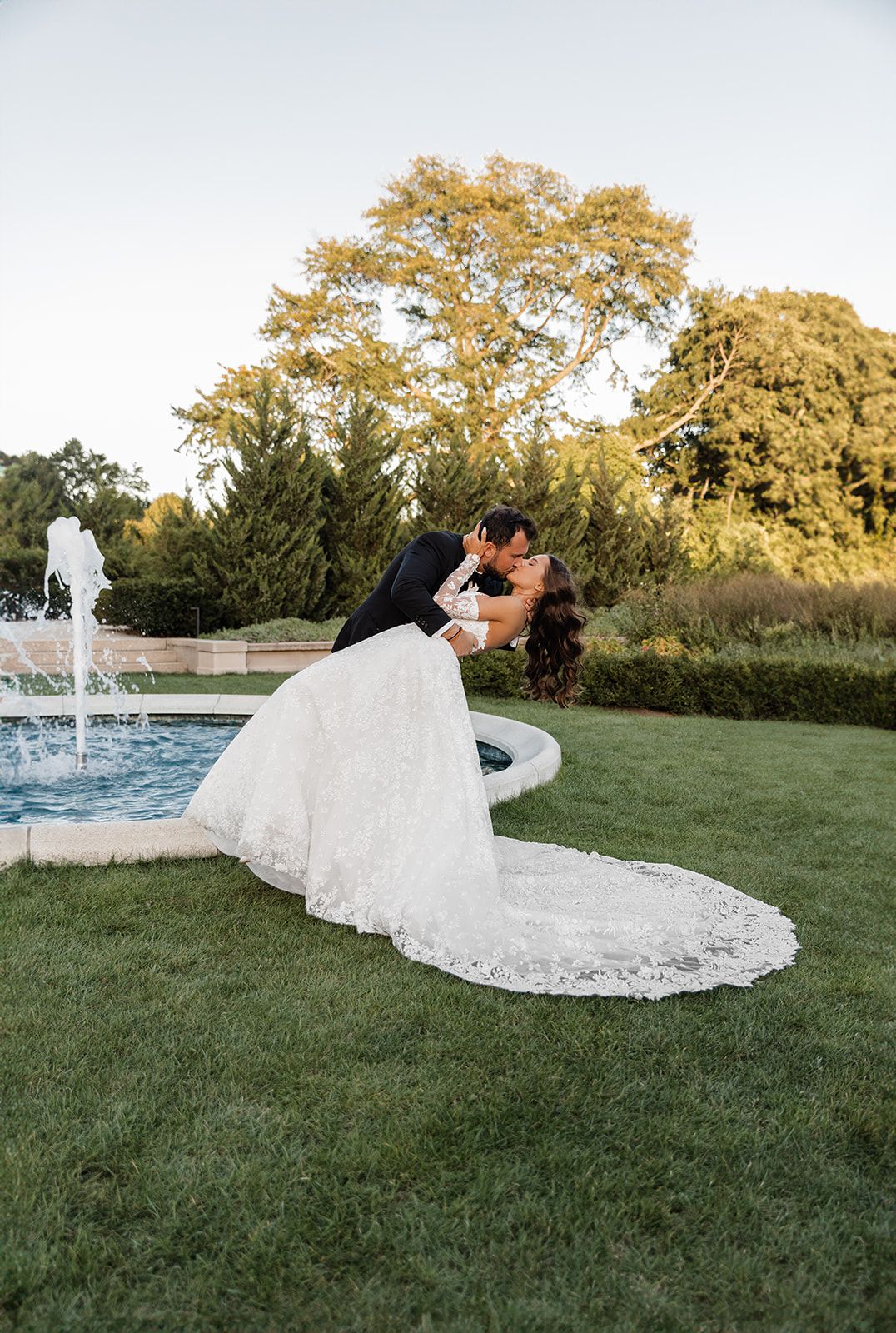 Bride and groom share a romantic dip kiss by a fountain in a lush garden, bride in lace gown.