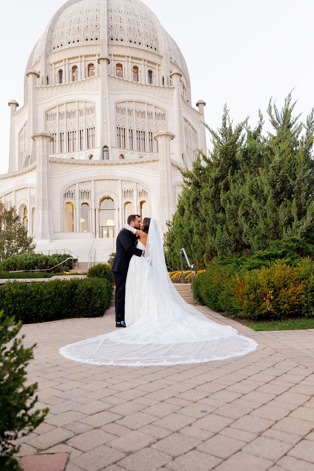 Bride and groom kissing in front of the Bahá'í Temple in Wilmette, Illinois, with a long cathedral veil spread on the ground.