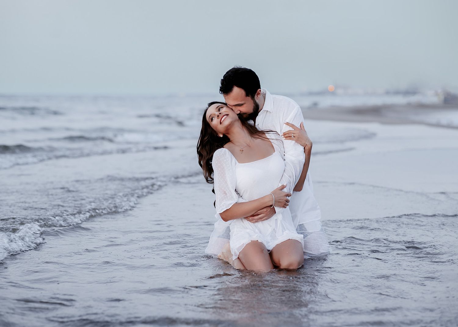 Romantic couple in white outfits embracing on a beach shore, waves gently surrounding them at dusk.
