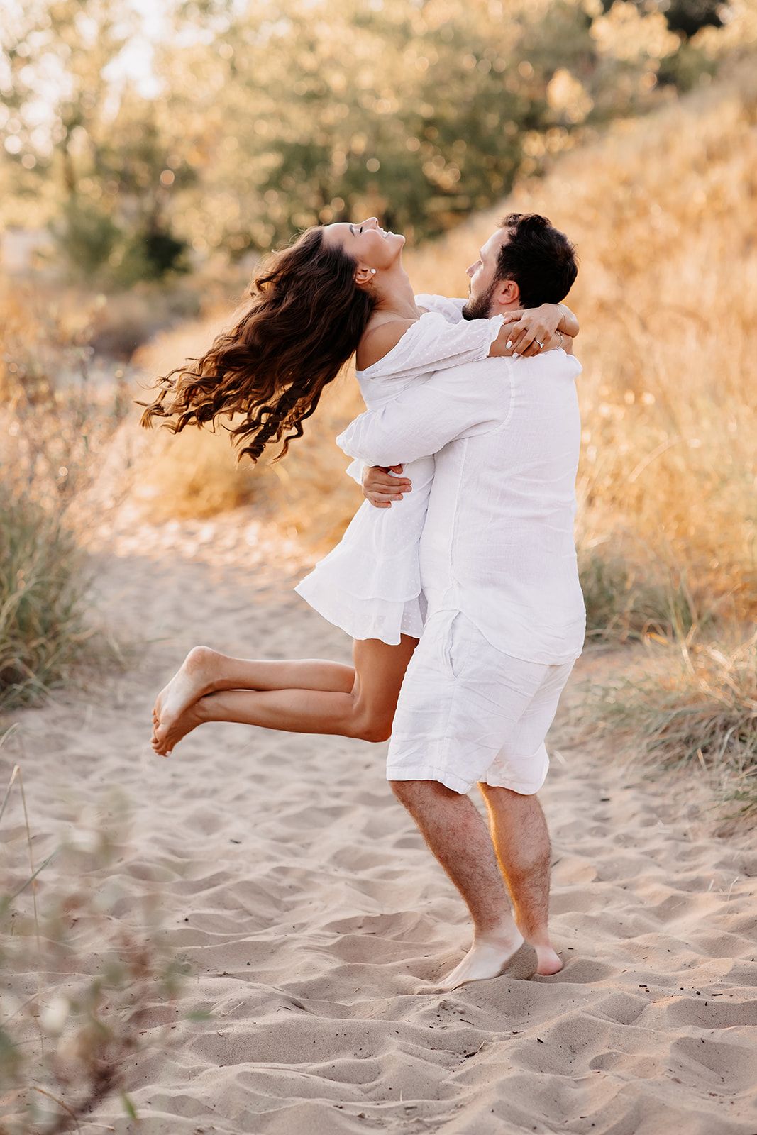 Man lifting smiling woman in white dress on sandy beach path with golden trees in background.