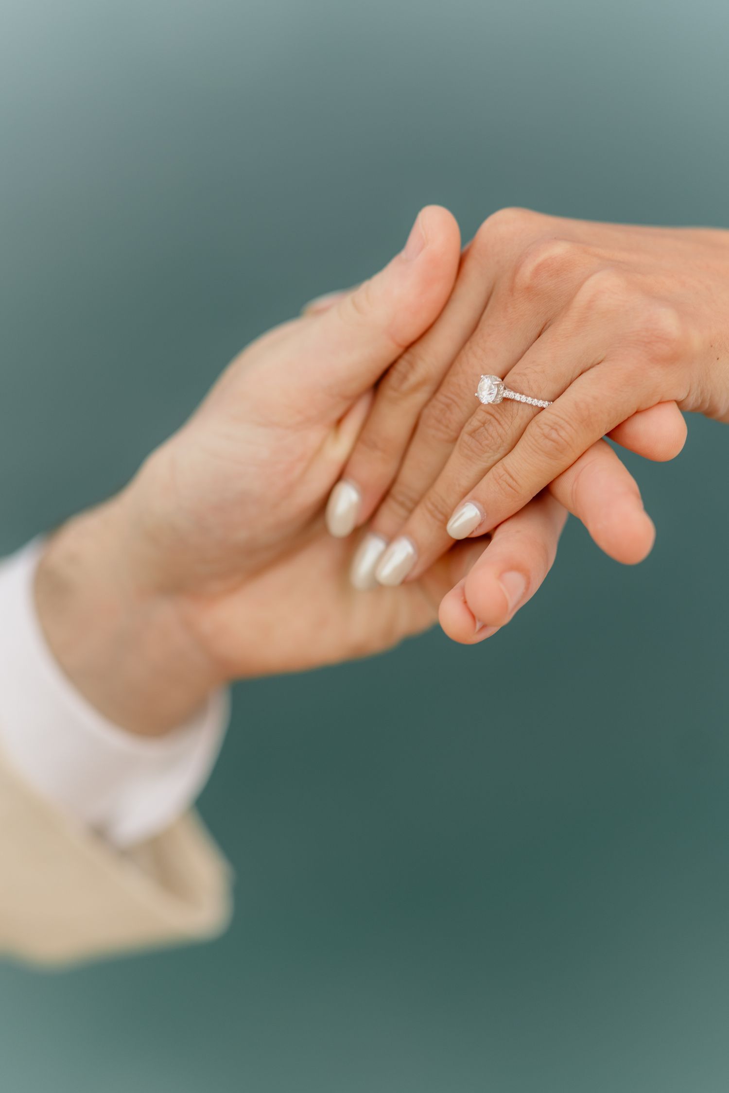 Two hands touching gently, one wearing a elegant diamond engagement ring with white manicured nails.