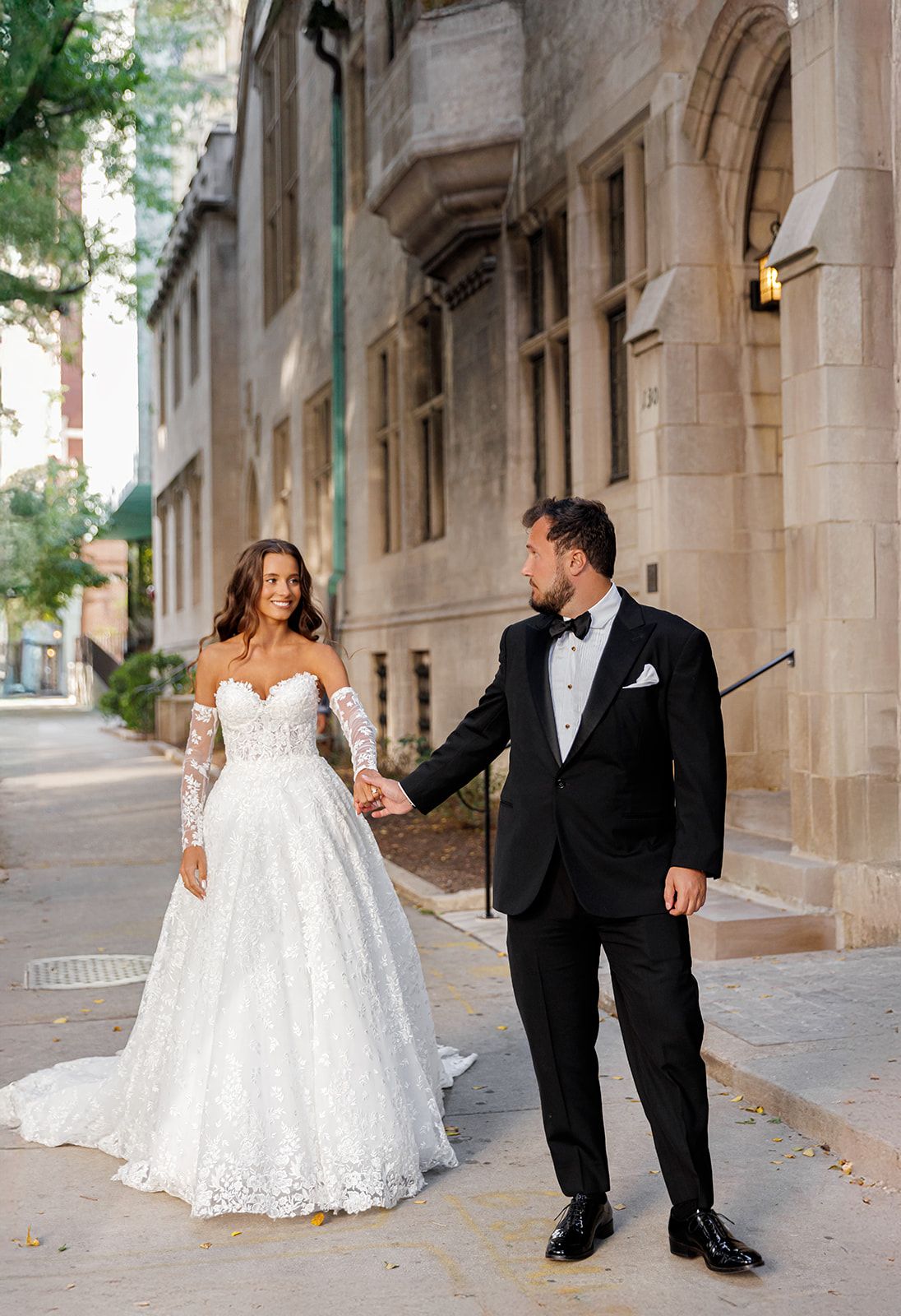 Bride in lace off-shoulder gown holds hands with groom in black tuxedo on a charming urban street.