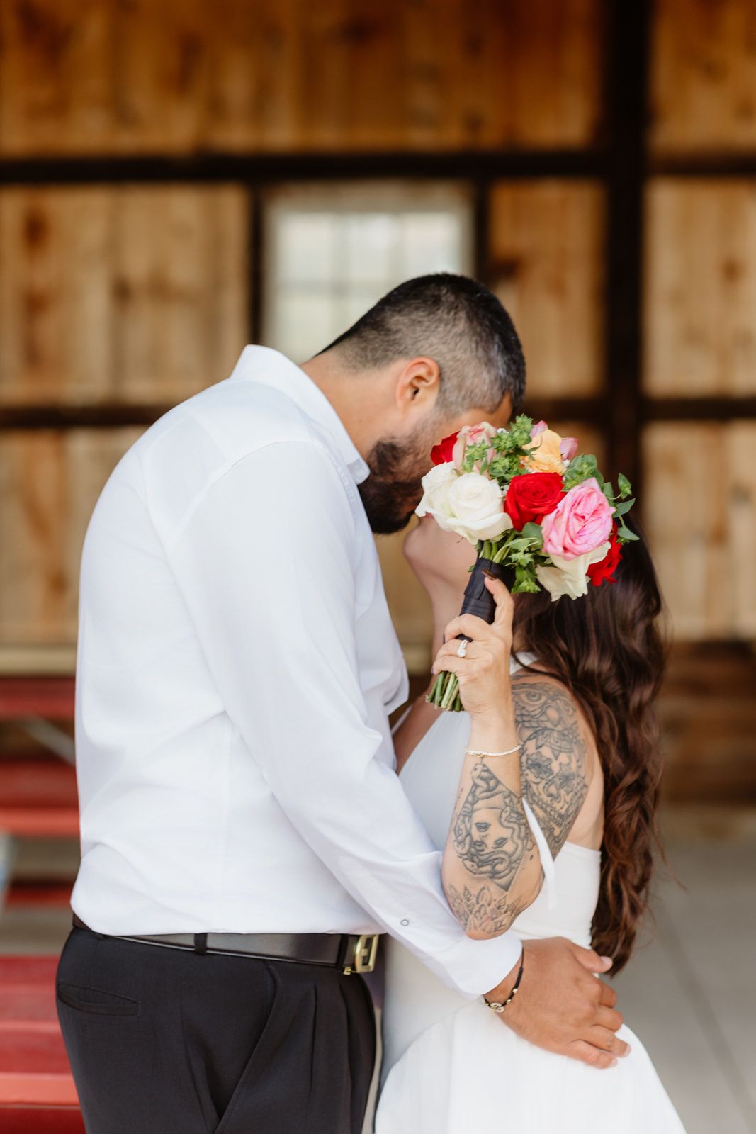 Bride and groom share a romantic kiss hidden behind a vibrant pink and red rose bouquet in a rustic barn setting.