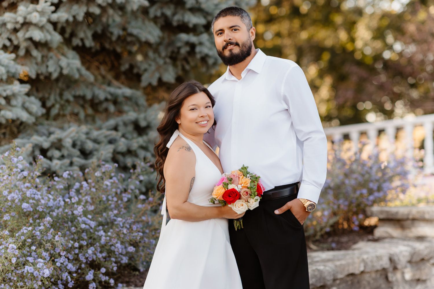 Smiling bride and groom pose together outdoors holding colorful bouquet, bride in white dress, groom in white shirt.