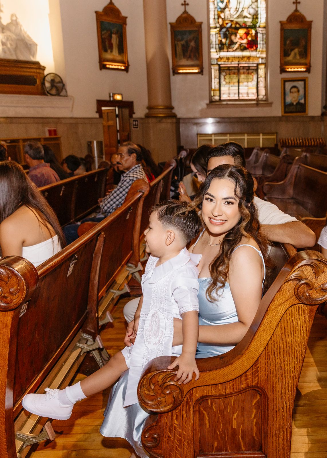 Woman and young child sitting in wooden church pews during a religious ceremony with stained glass windows.