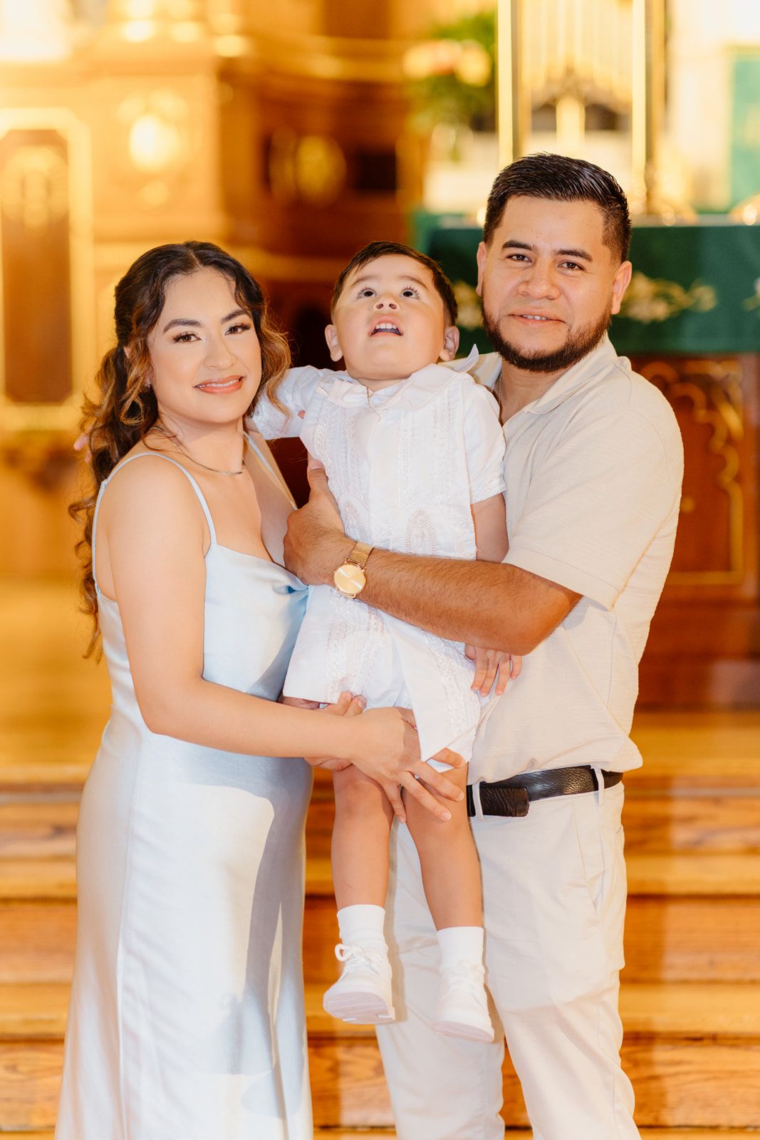 A young Hispanic family poses in a church, parents holding their toddler in white baptism outfit.