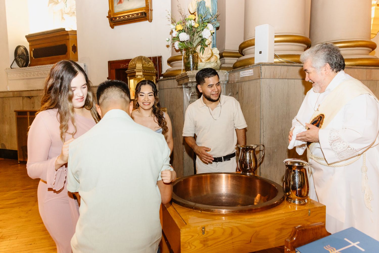 Family and priest gathered around copper baptismal font before Catholic baptism ceremony in church.