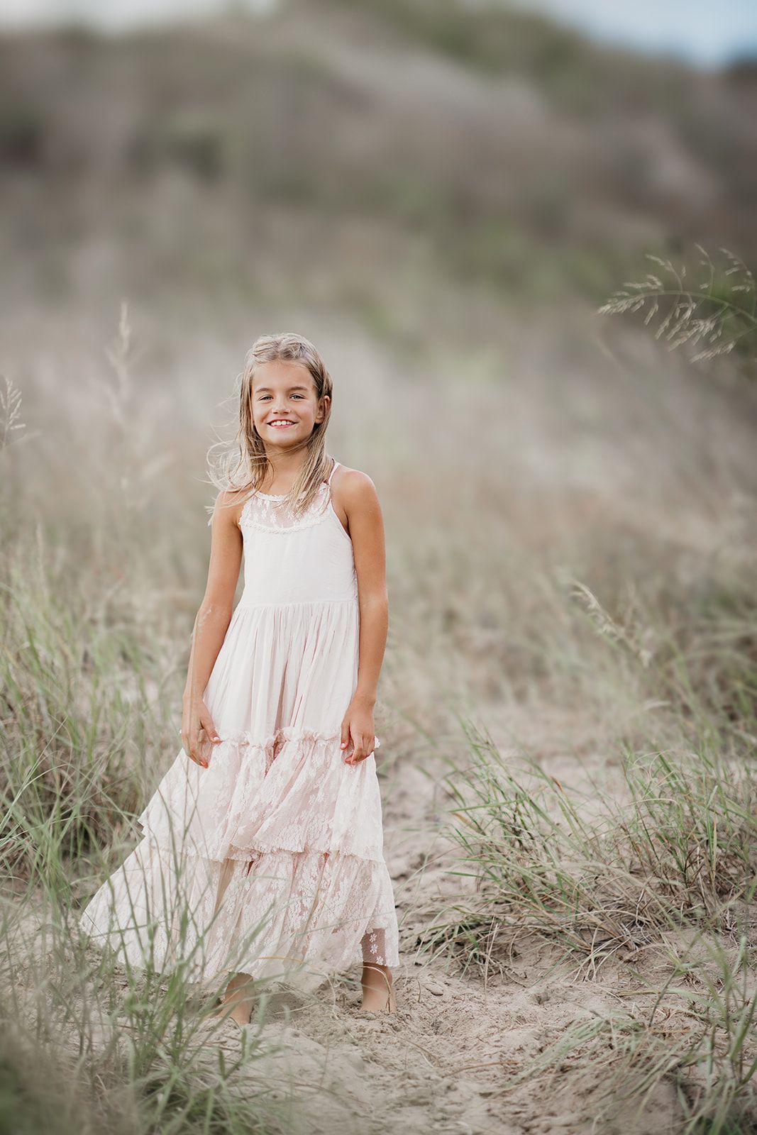 A smiling young girl in a flowing blush pink tiered maxi dress stands in a sandy field with tall grass.