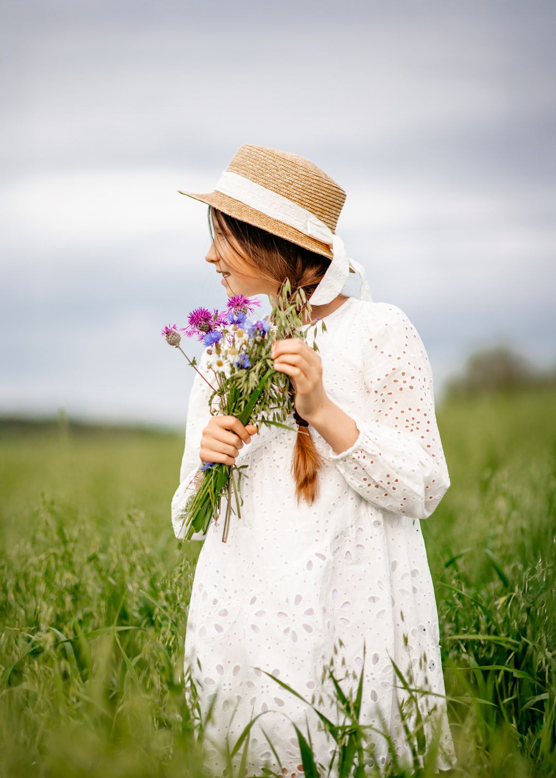 A young woman in a white eyelet dress and straw hat holds wildflowers in a lush green field.