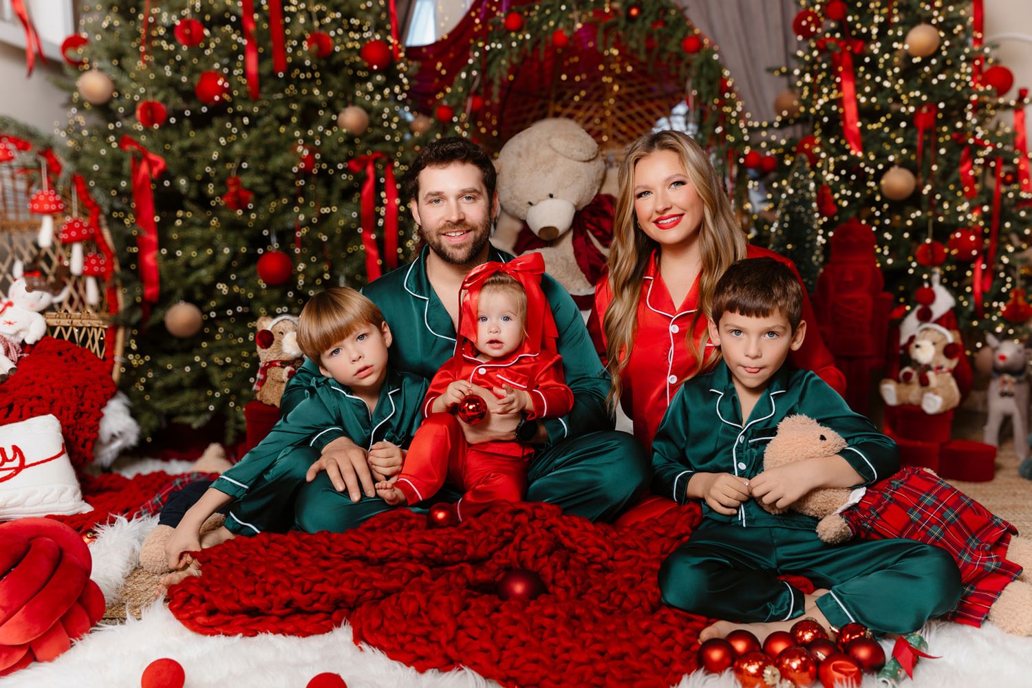 Family of five in matching Christmas pajamas posing by decorated tree with red ornaments and teddy bear.