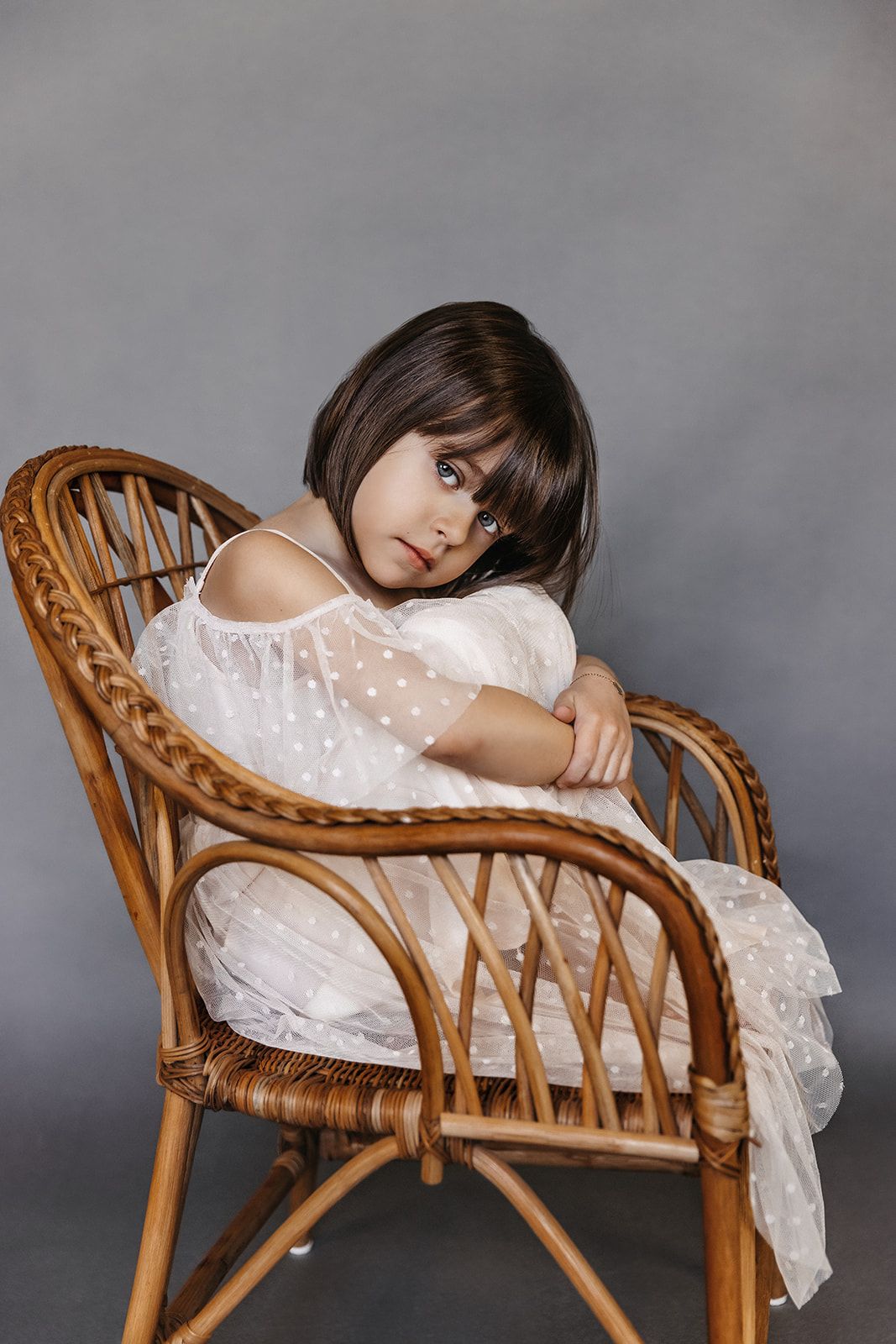 Young girl in white polka dot dress sitting in wicker chair against gray background.
