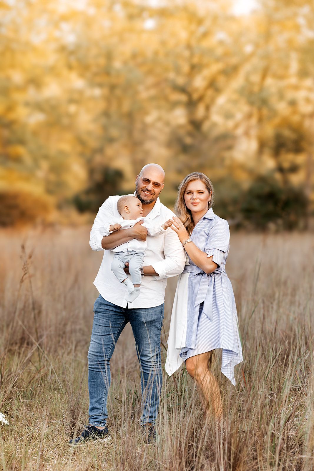 A couple stands in a golden autumn field, the man holding their baby while the woman leans in closely.