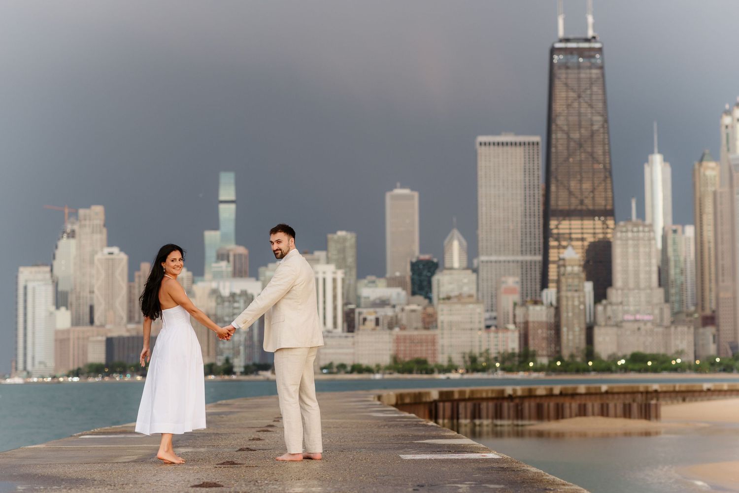 Engaged couple holds hands barefoot on pier with iconic Chicago skyline and John Hancock tower behind them.