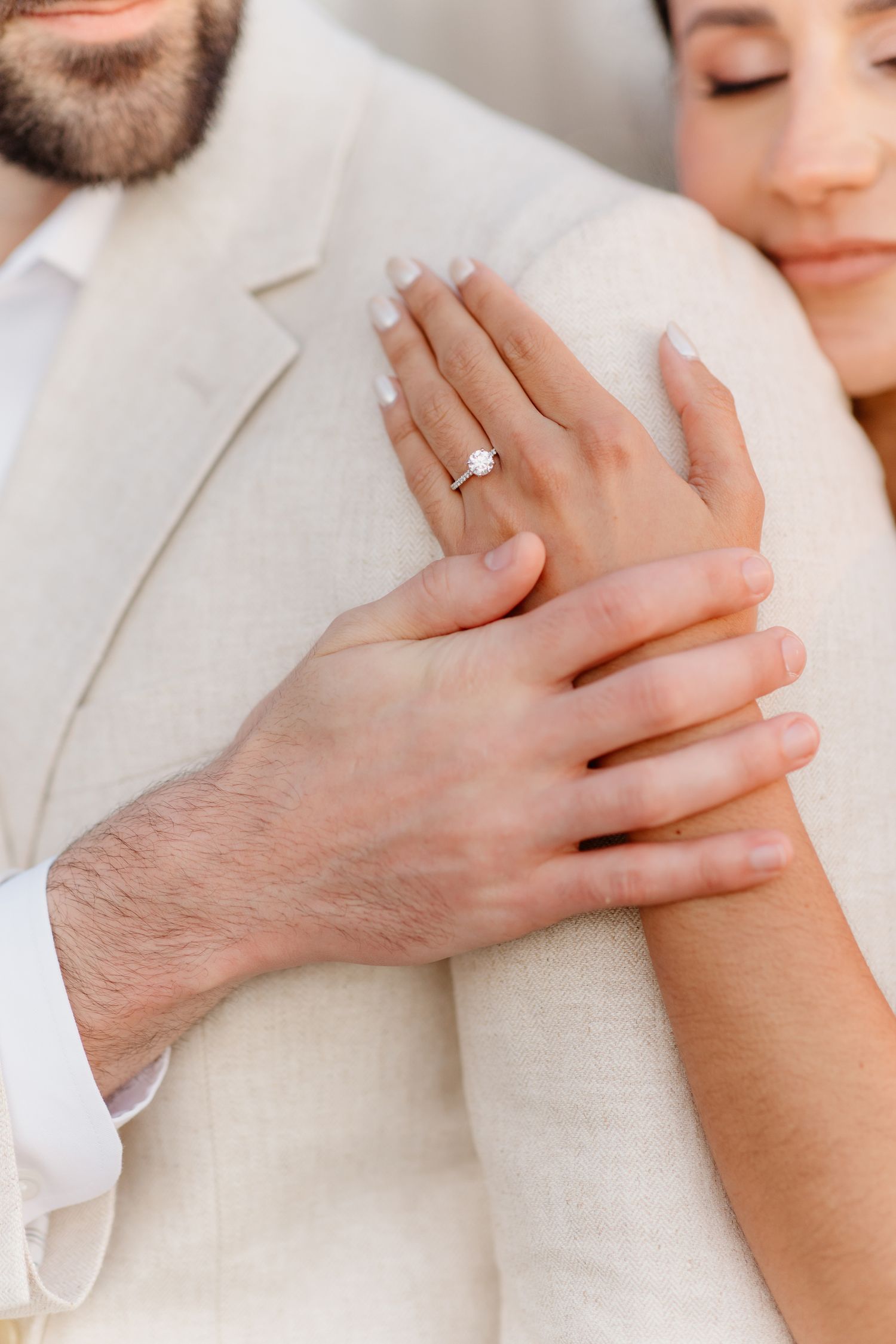 Close-up of bride's hand displaying diamond engagement ring while resting on groom's beige linen suit.