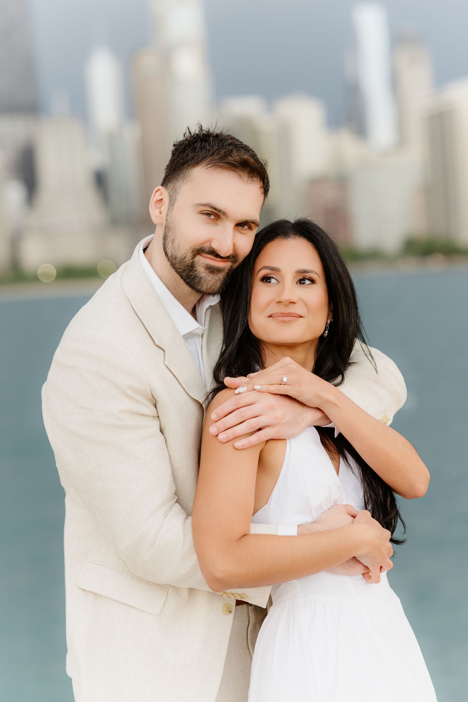 Couple embracing by Chicago lakefront, man in beige suit hugging woman in white dress with city skyline behind.