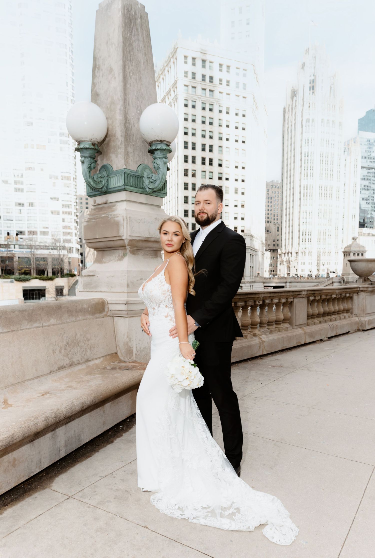 Bride in lace mermaid gown and groom pose on Chicago bridge with city skyline backdrop.