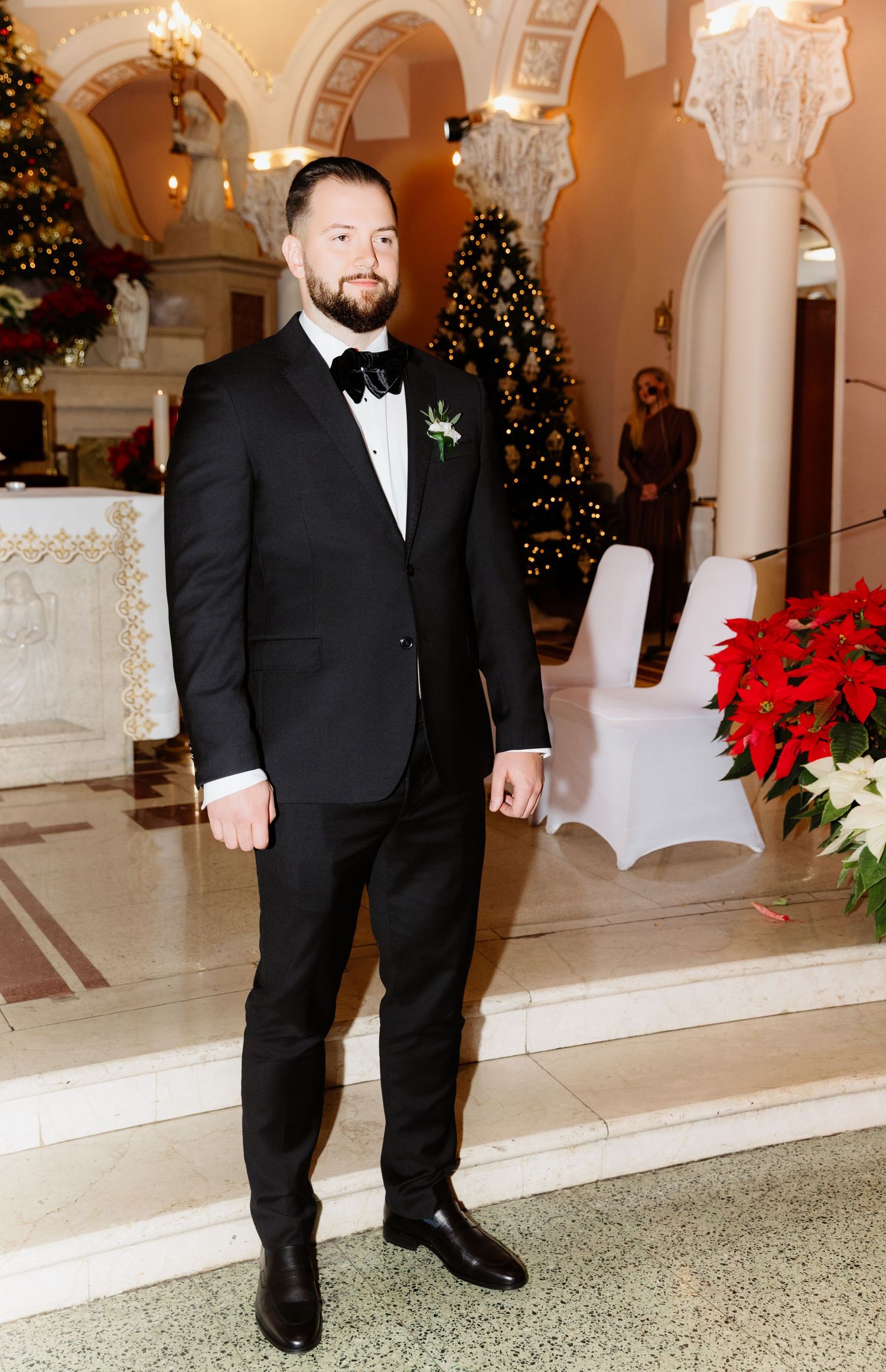 A groom in a black tuxedo with boutonniere poses near poinsettias and a Christmas tree in a church.