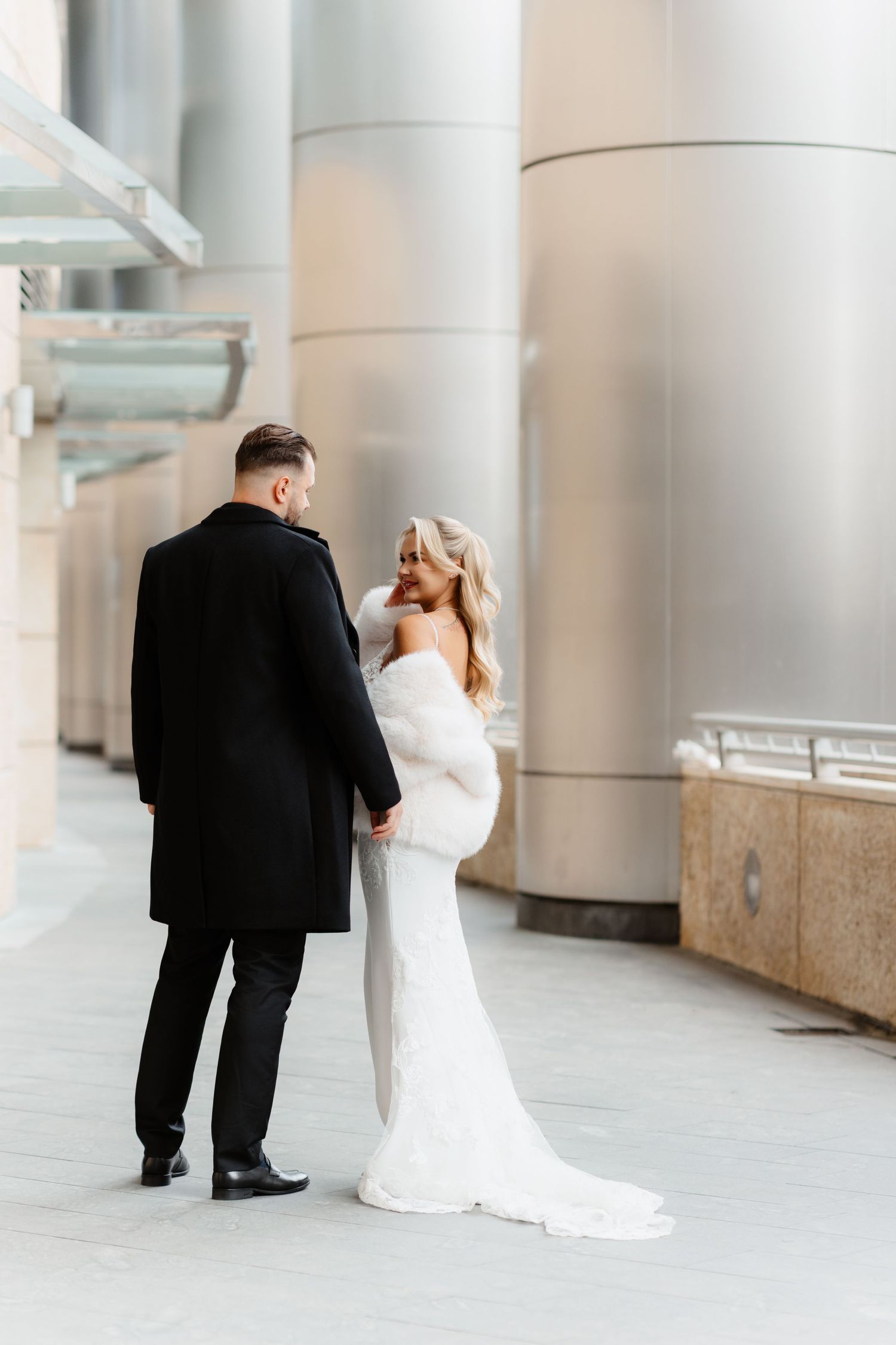 Bride in white fur wrap smiles at groom in black coat beside large silver columns outdoors.
