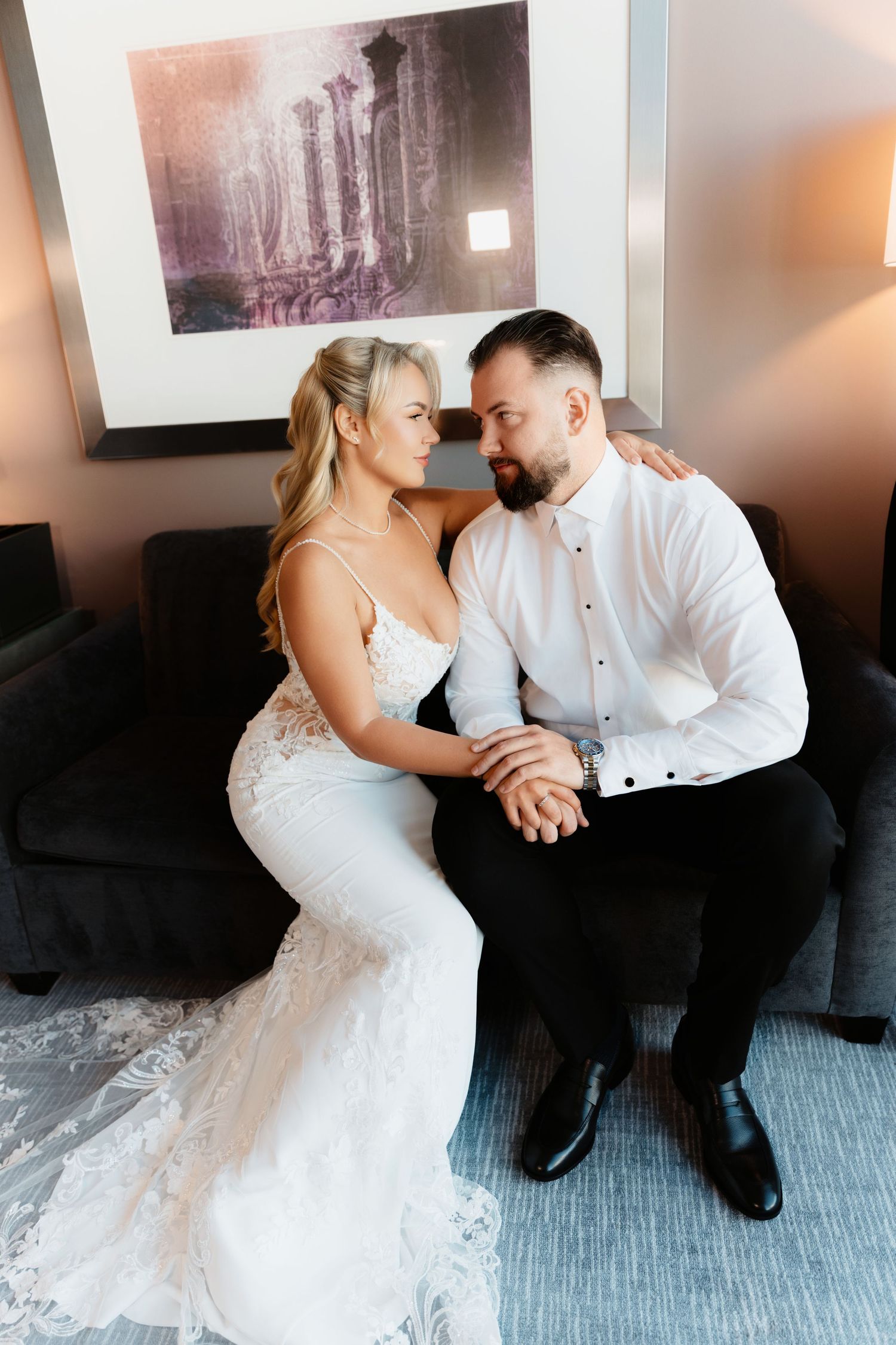 Bride in lace wedding dress and bearded groom sit close together on dark sofa, foreheads touching.