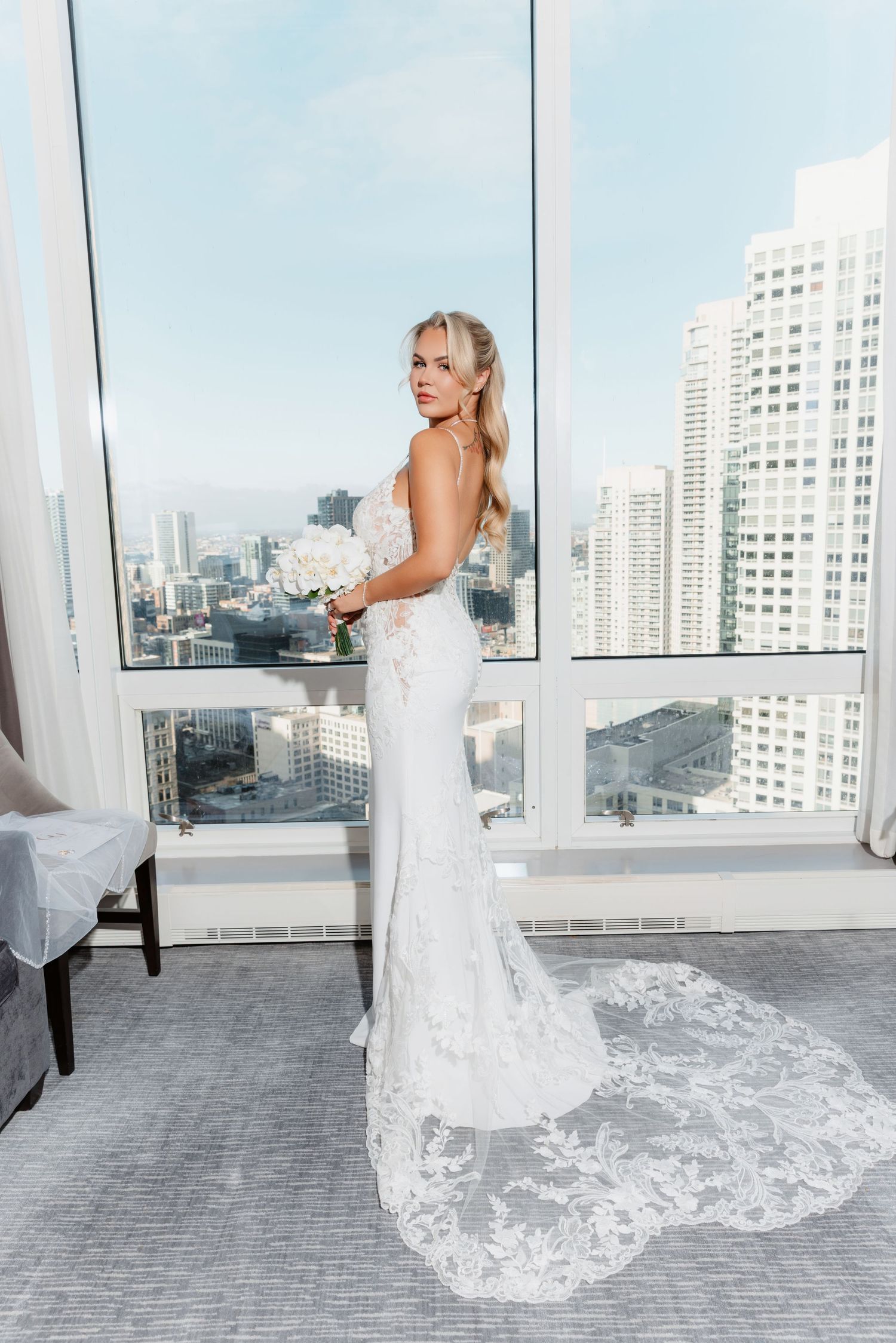Bride in backless lace mermaid gown holding white bouquet poses before floor-to-ceiling city view windows.