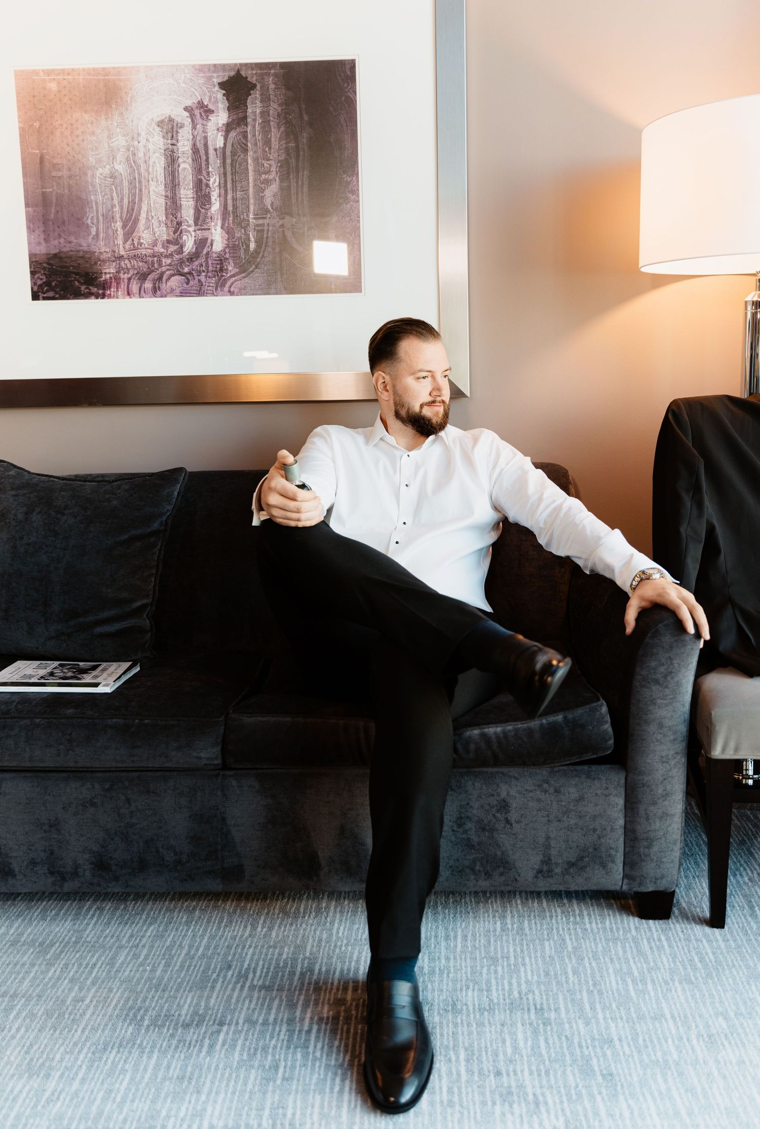 Groom in white shirt relaxes on gray sofa holding drink during wedding day preparations.
