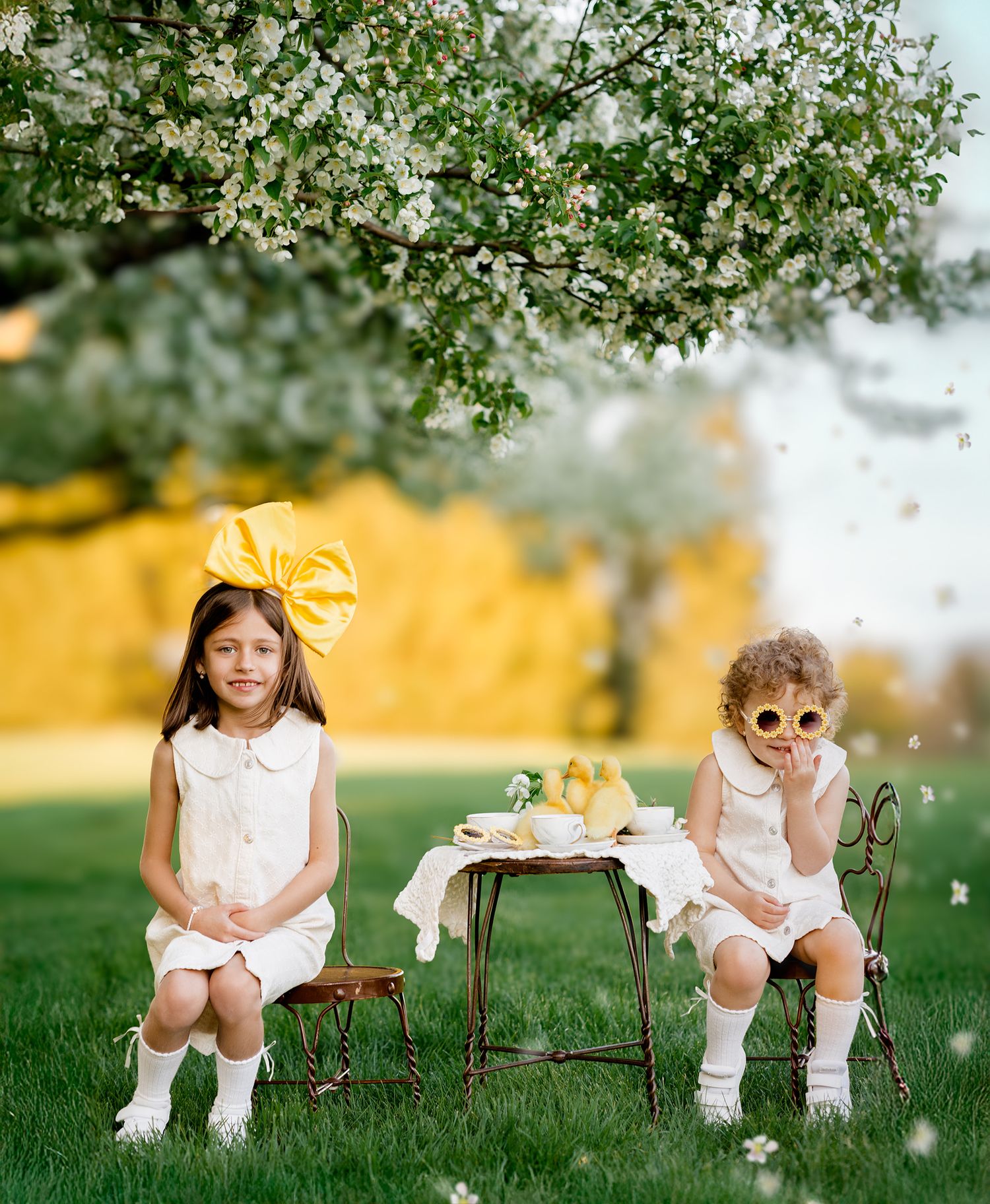 Two young girls in white dresses enjoy a tea party outdoors under a blooming spring tree on lush green grass.